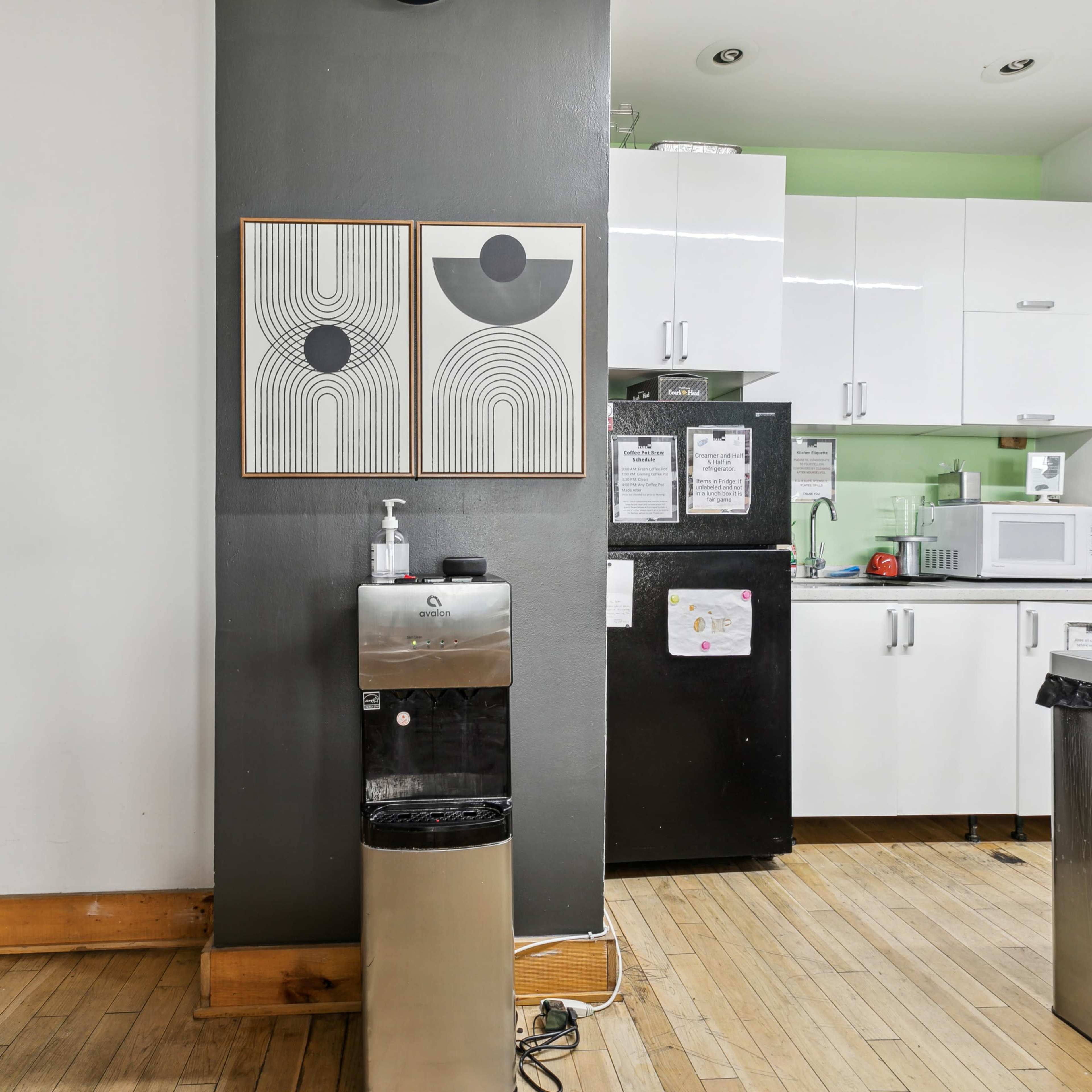 A kitchen area with a water dispenser, artwork on the wall, and various kitchen appliances, including a microwave and refrigerator.