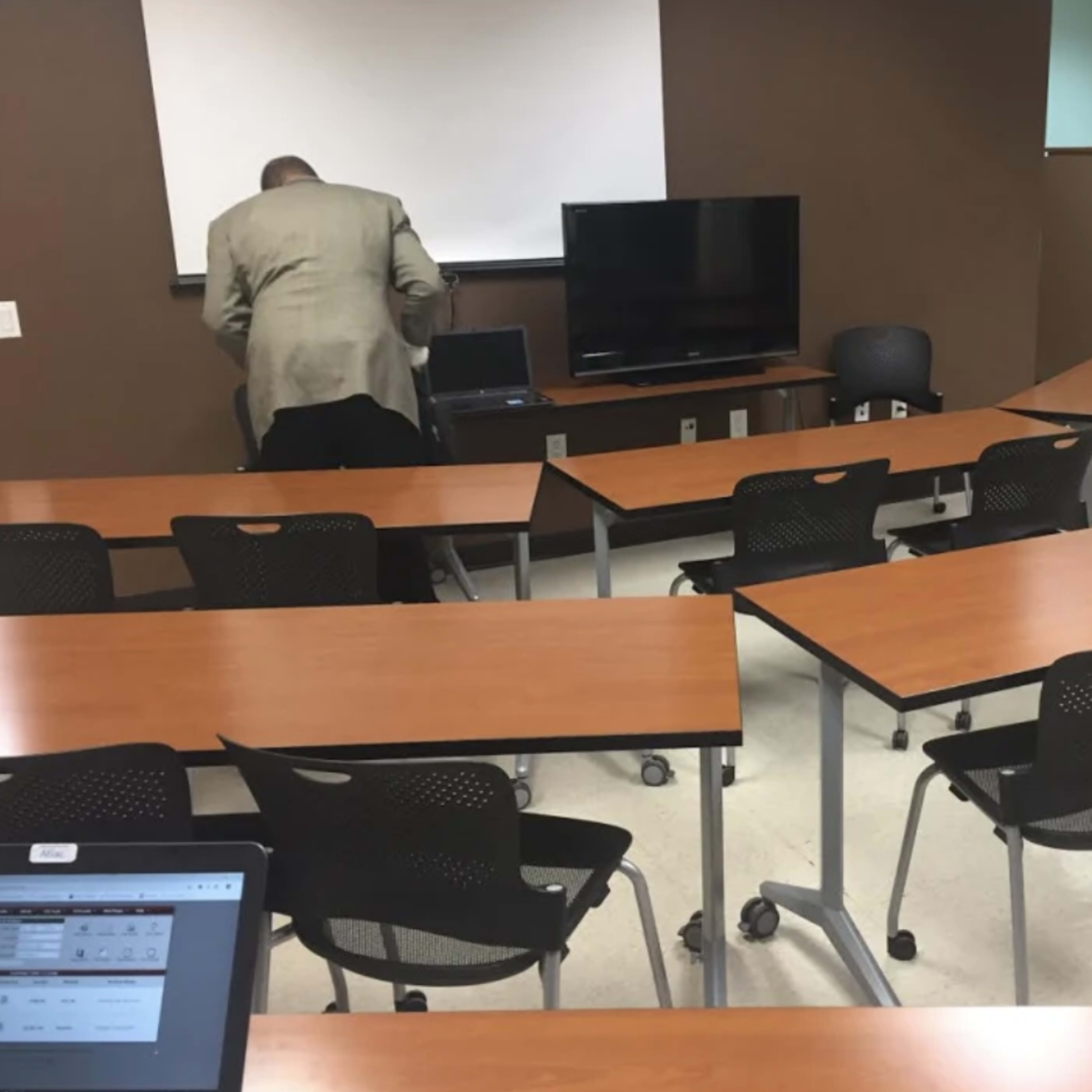 A man in a suit stands near a laptop in an empty classroom with movable tables and chairs arranged in a U-shape.