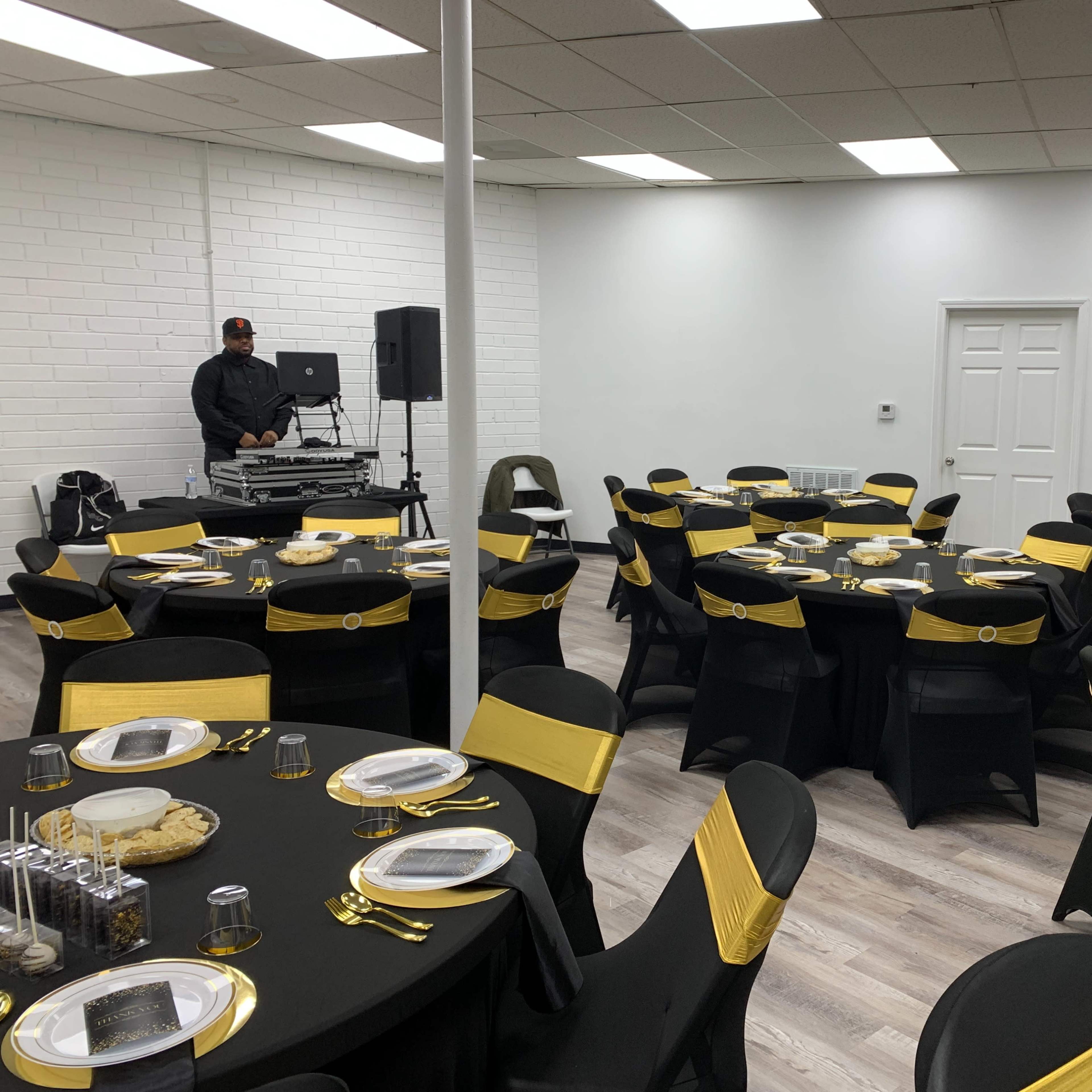 A large event space features black tables with yellow accents, each set with plates and utensils, while a DJ stands in the corner.
