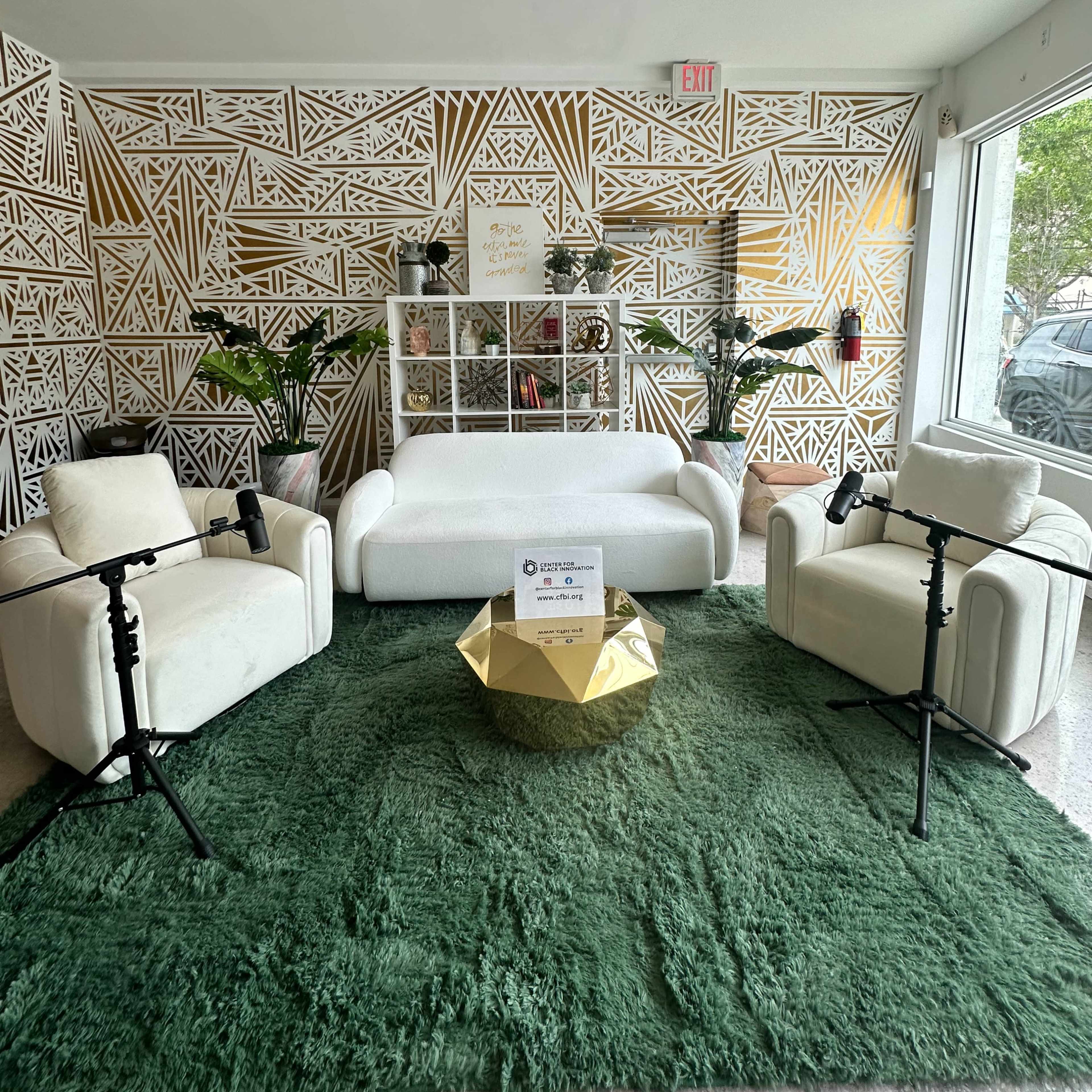 A modern lounge area featuring a white sofa, two white chairs, a green rug, and a gold coffee table, with a decorative wall design in the background.