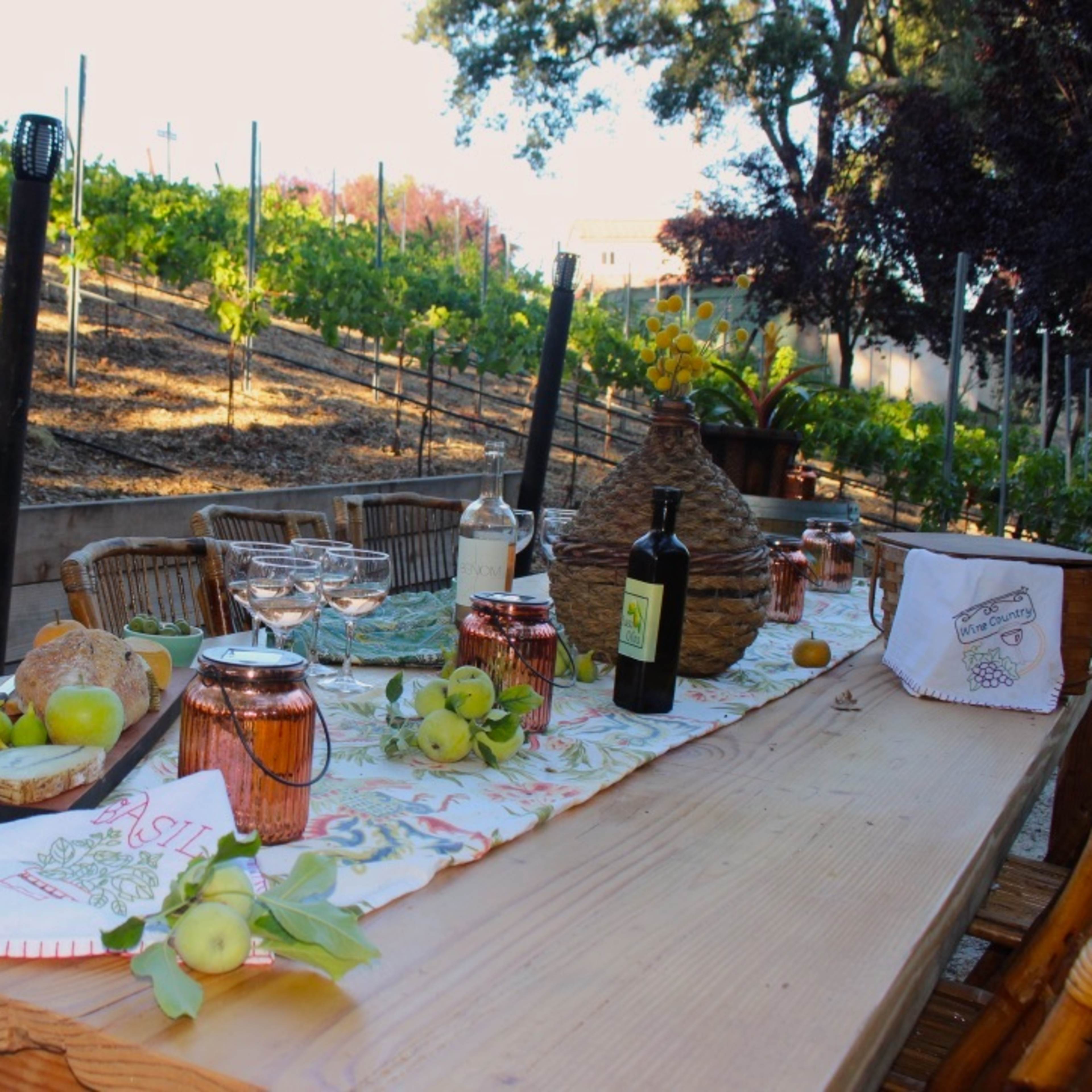 A rustic wooden table is set with food and drinks, surrounded by a vineyard on a hillside.