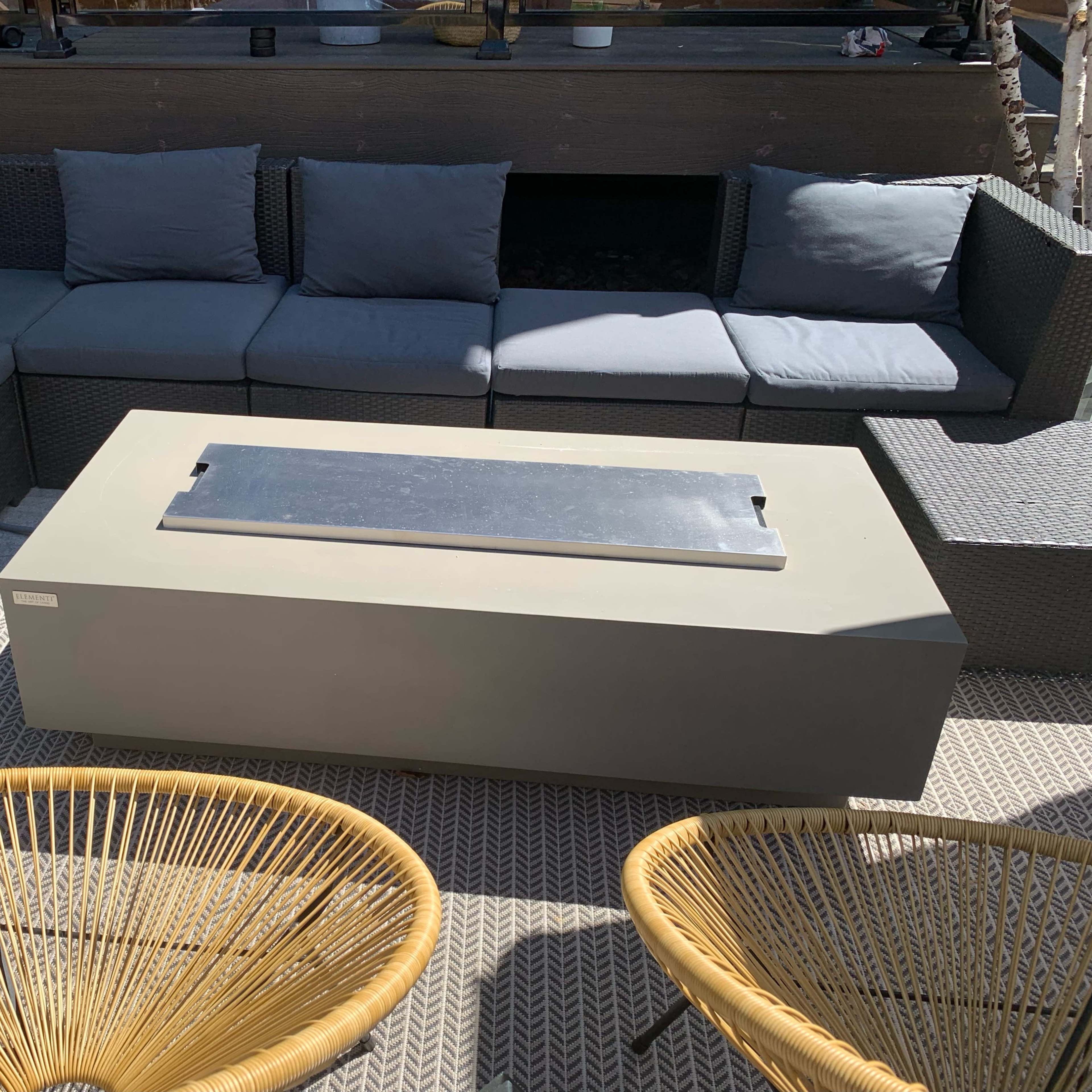 A modern outdoor seating area features a gray sofa arrangement and a rectangular fire table surrounded by two woven chairs.