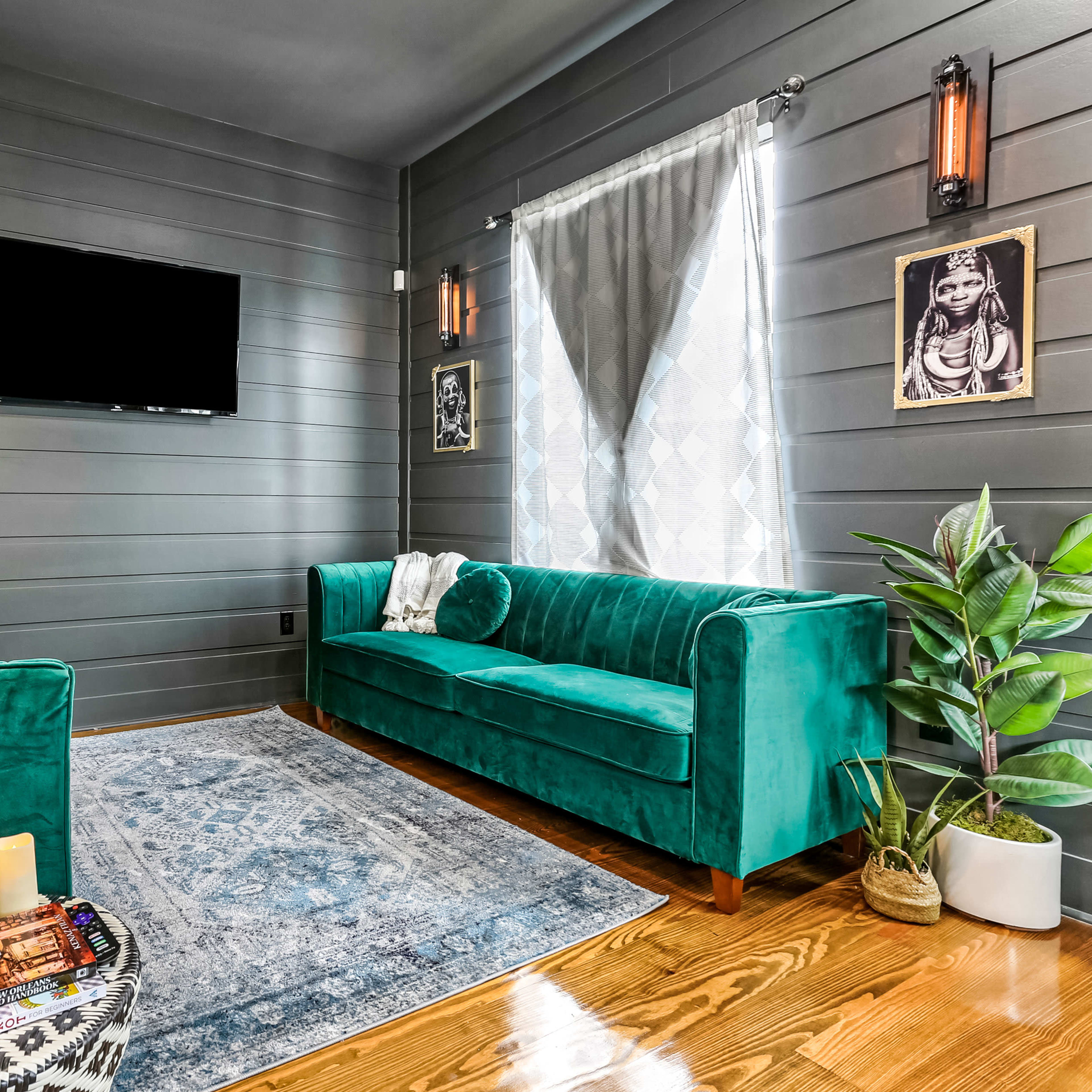 The room features two teal sofas, a patterned rug, a television mounted on the wall, and a large window with sheer curtains.