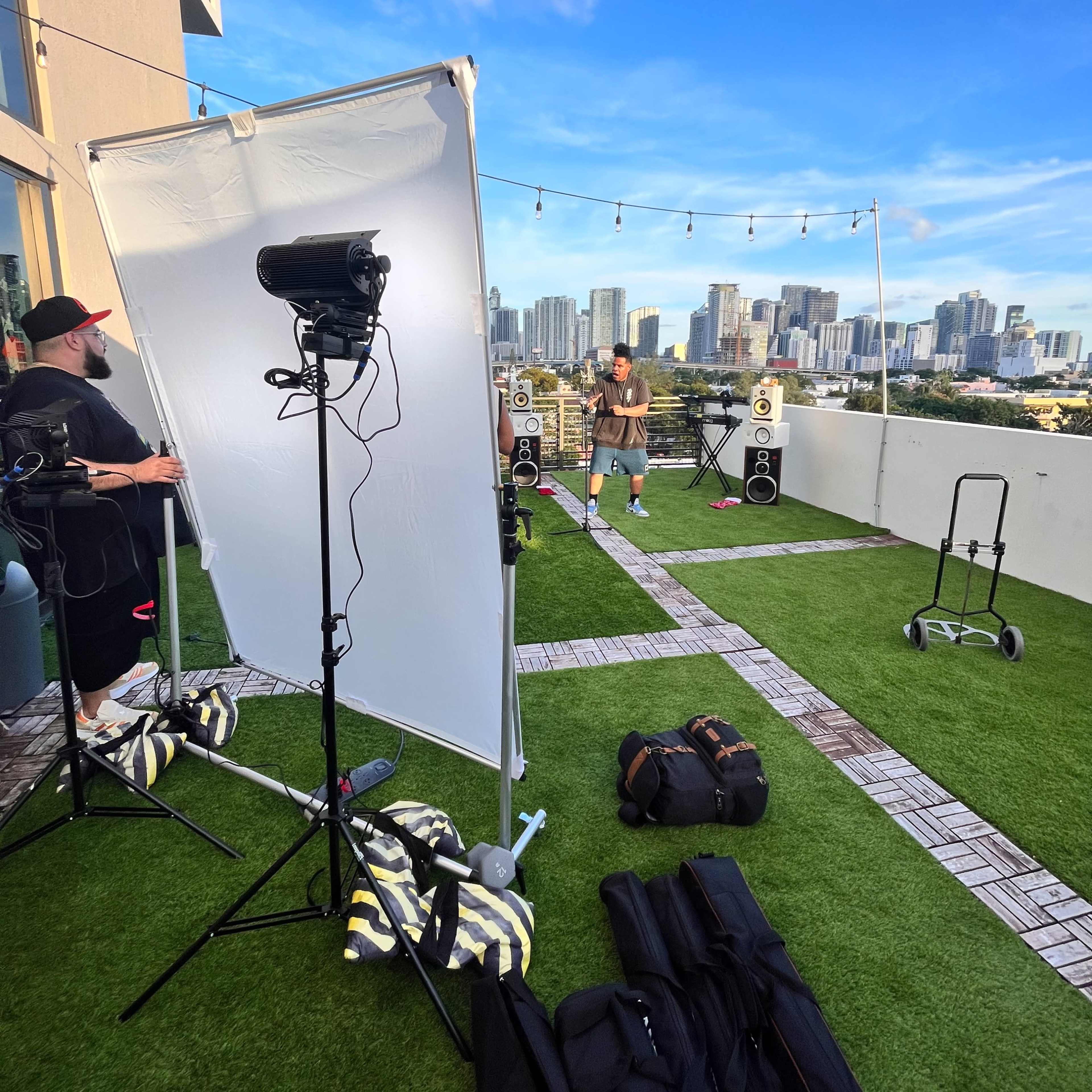 A man stands on a rooftop patio setting with city skyscrapers in the background, while two crew members adjust lighting equipment and speakers.