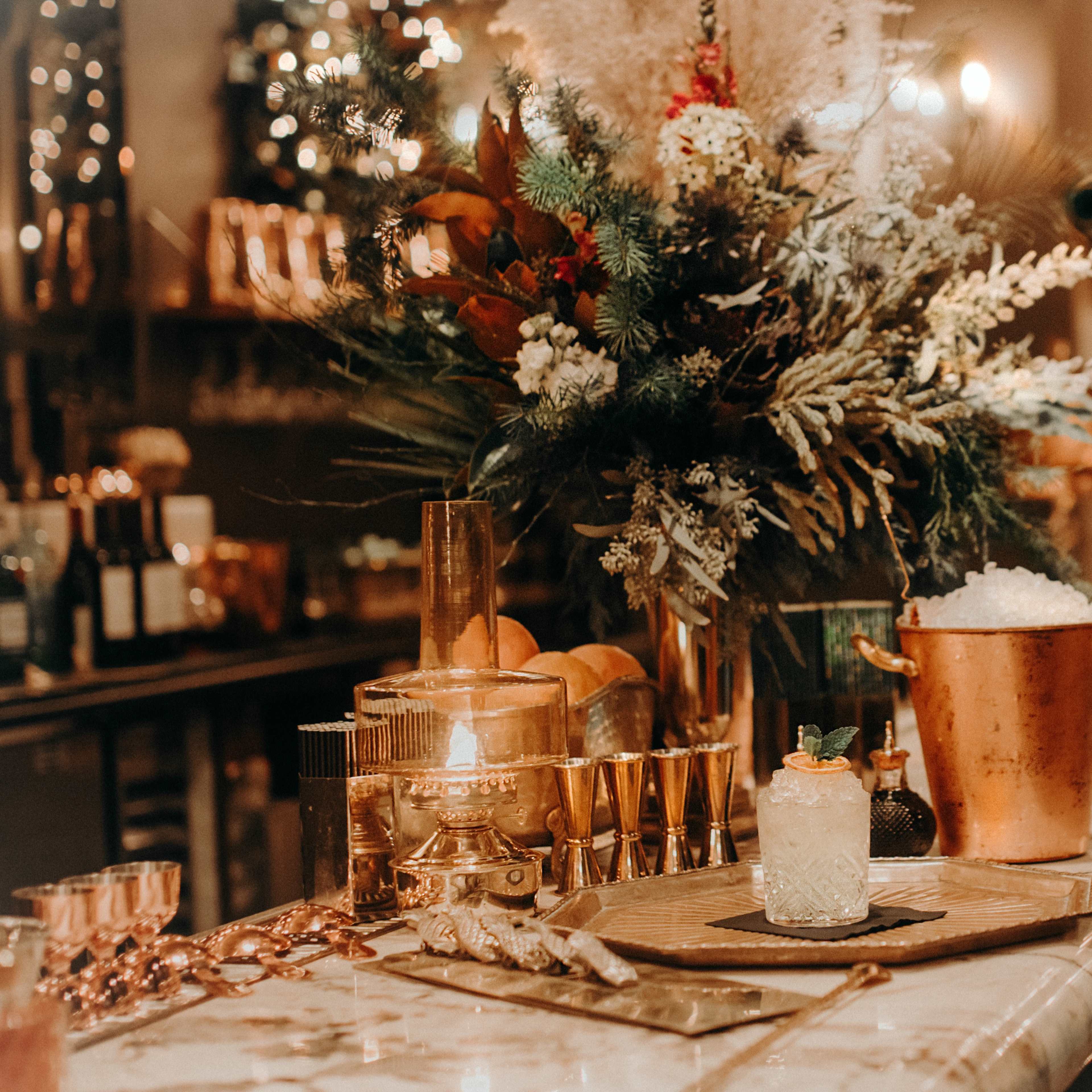 A beautifully arranged bar countertop with a large floral centerpiece, an ice bucket, cocktail glasses, and a decorated drink on a tray.