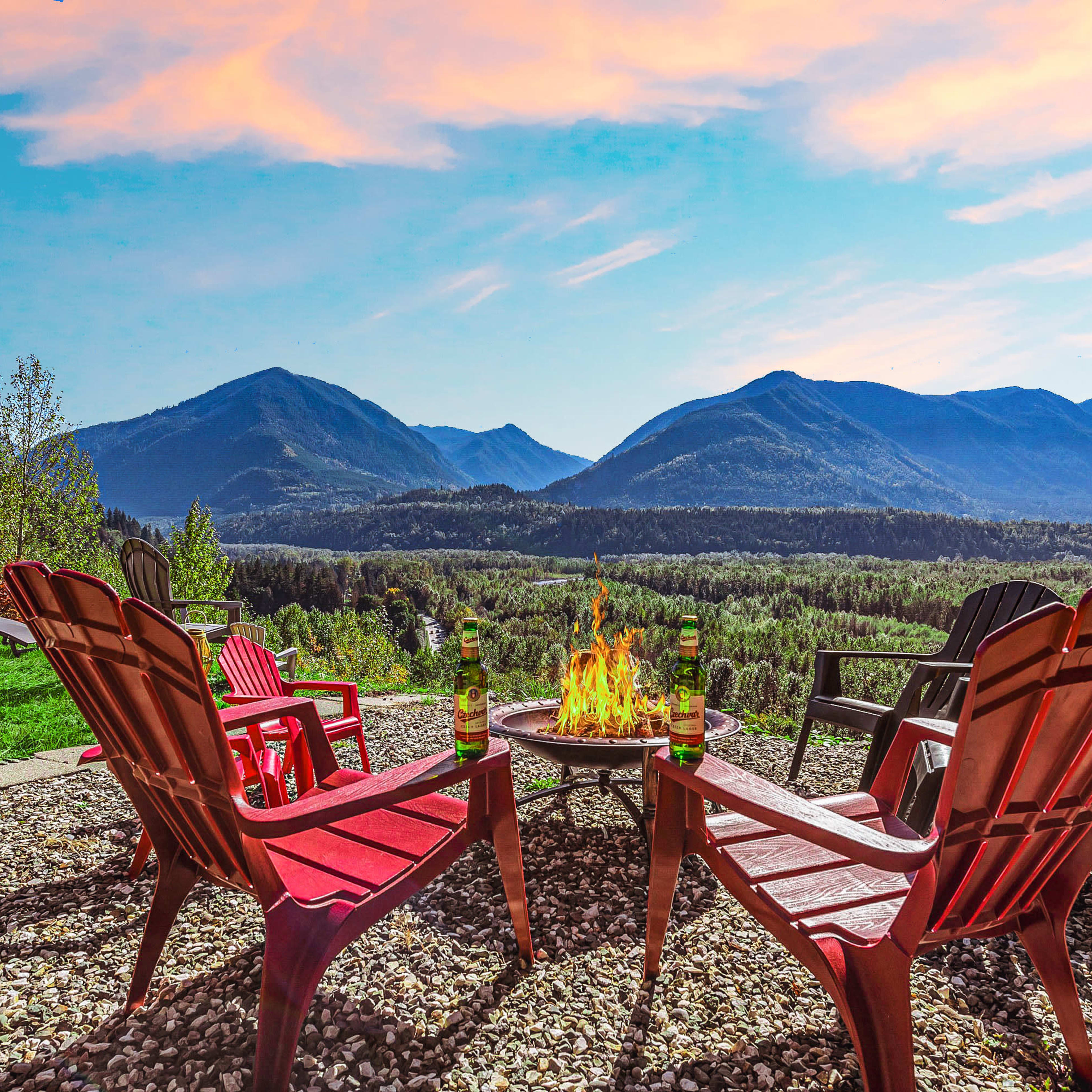 A circular arrangement of red Adirondack chairs surrounds a fire pit, with mountains and a colorful sky in the background.