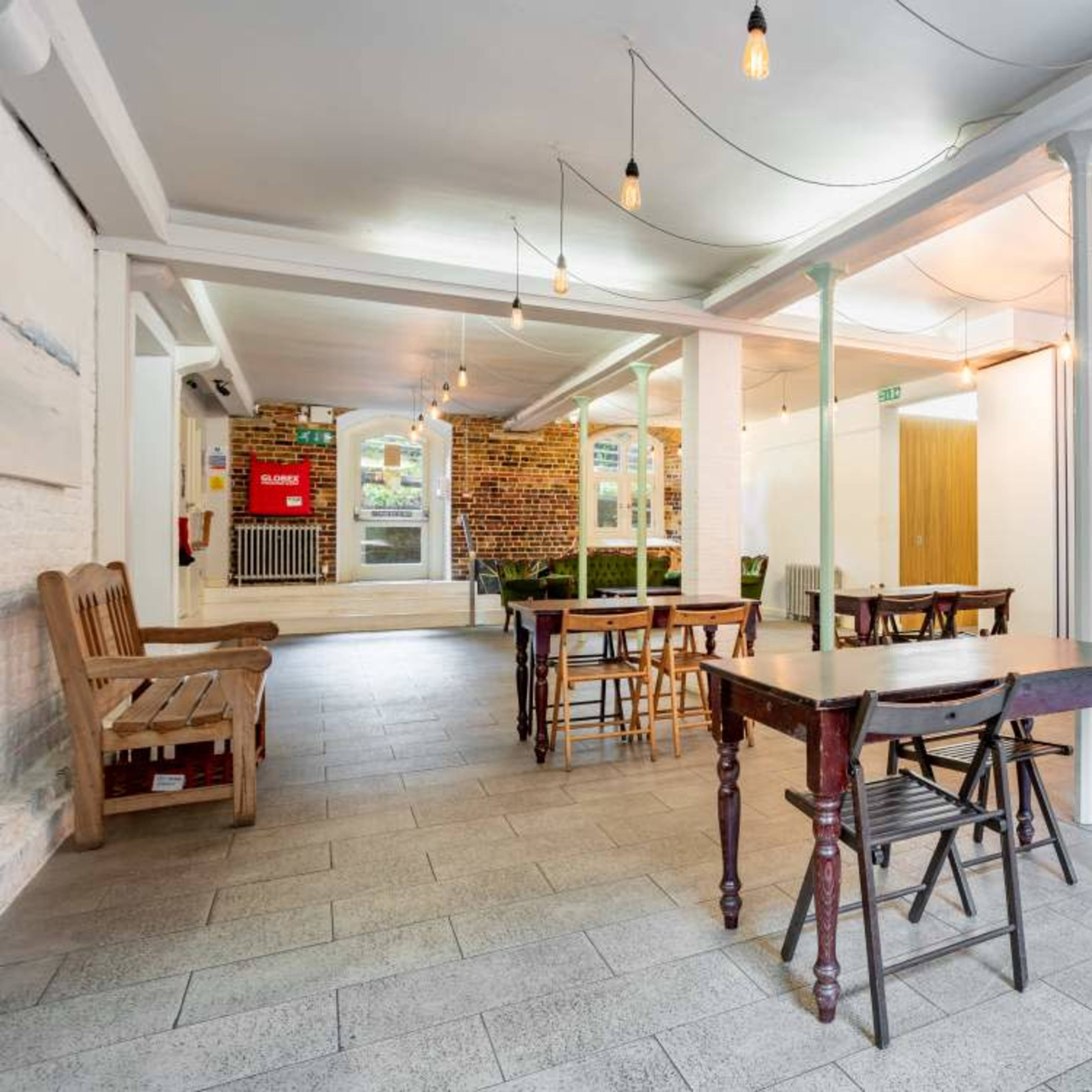 A spacious, well-lit interior with wooden tables and chairs arranged around a tiled floor, featuring hanging light bulbs and a few plants in the background.