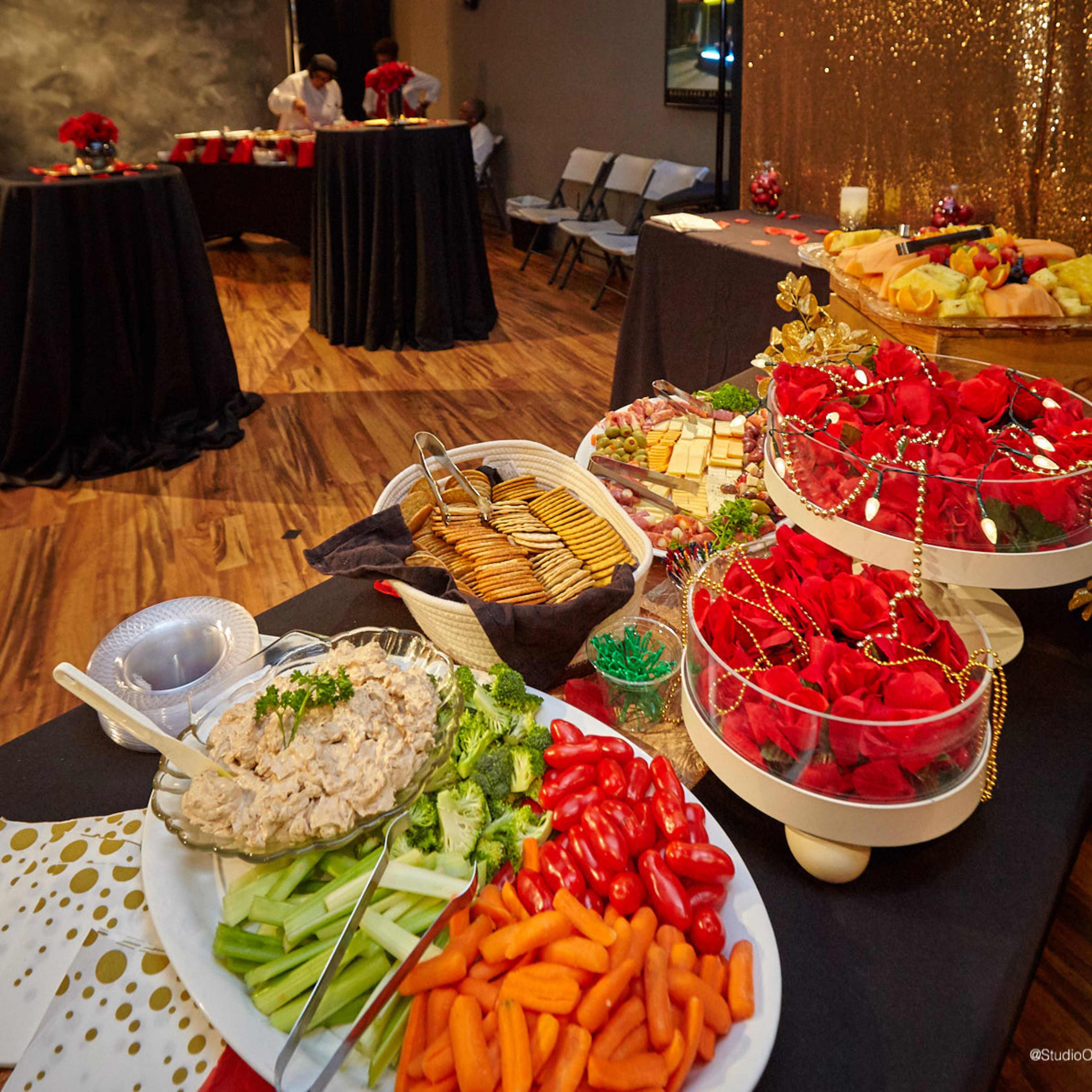 A buffet table is set with a variety of snacks, including a platter of vegetables, a bowl of dip, crackers, a cheese tray, and an arrangement of roses.