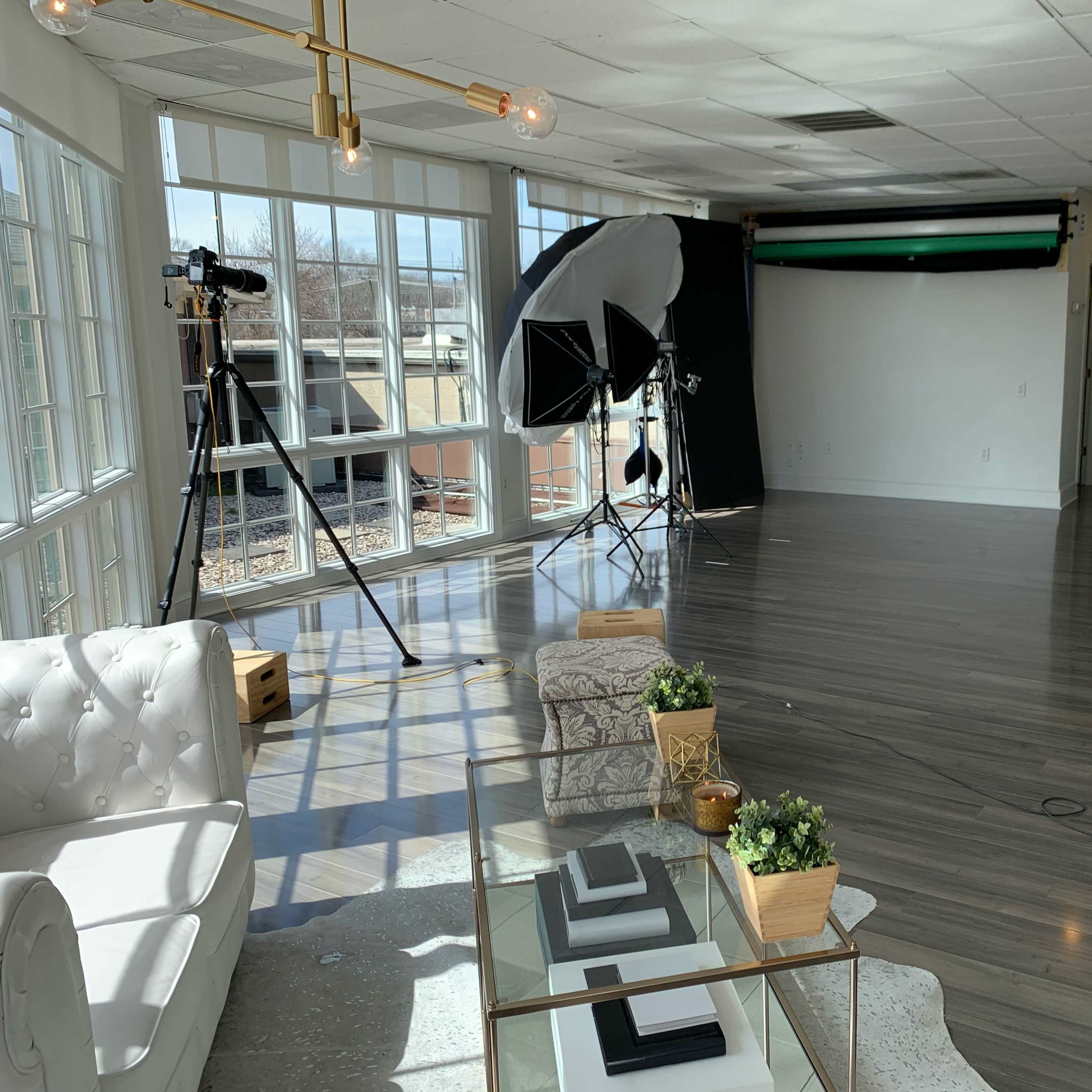 A well-lit studio space featuring a comfortable seating area, photography equipment, and a backdrop setup against a wall of windows.