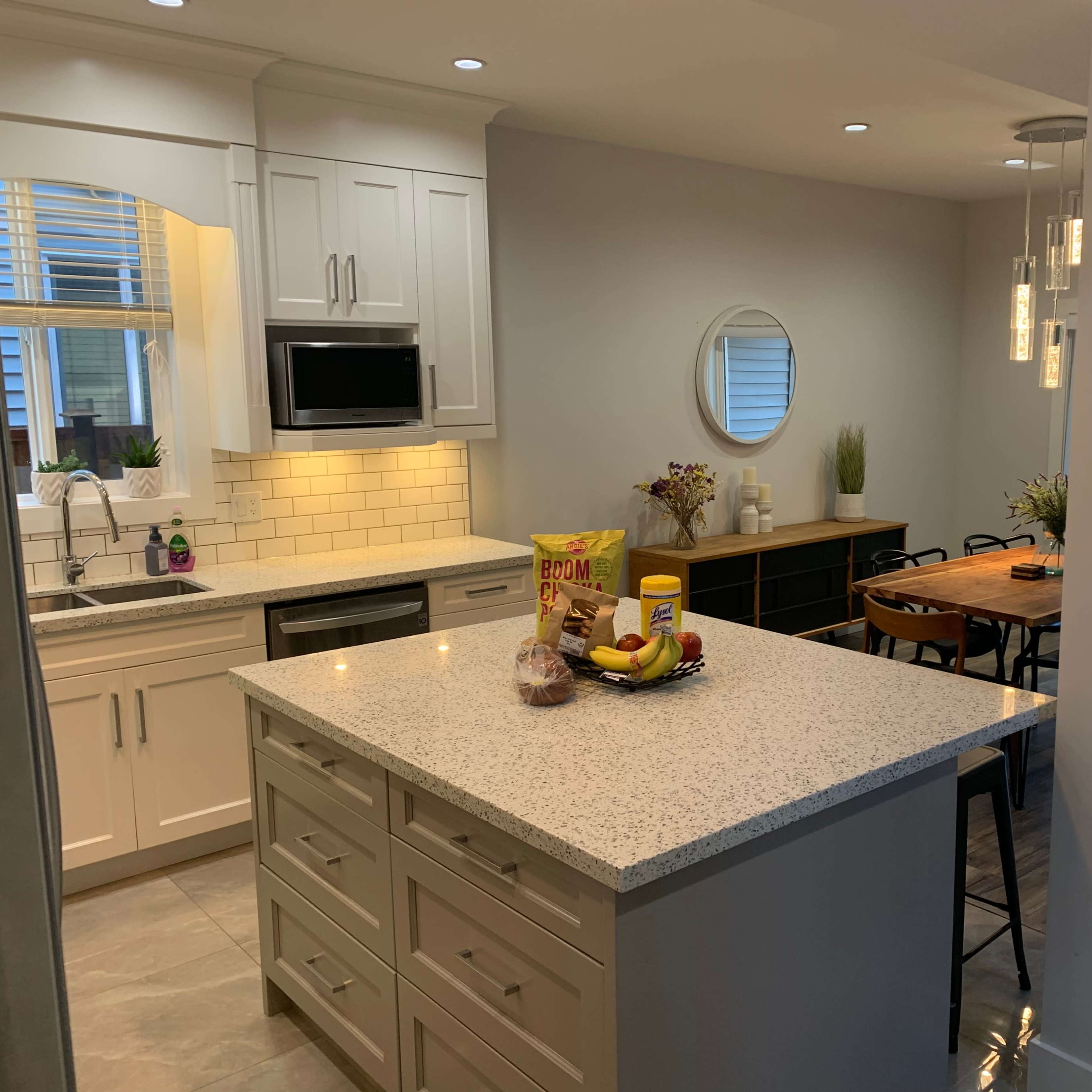 A modern kitchen with a central island, featuring white cabinetry, a stainless steel refrigerator, and a dining area with wooden furniture and large windows.