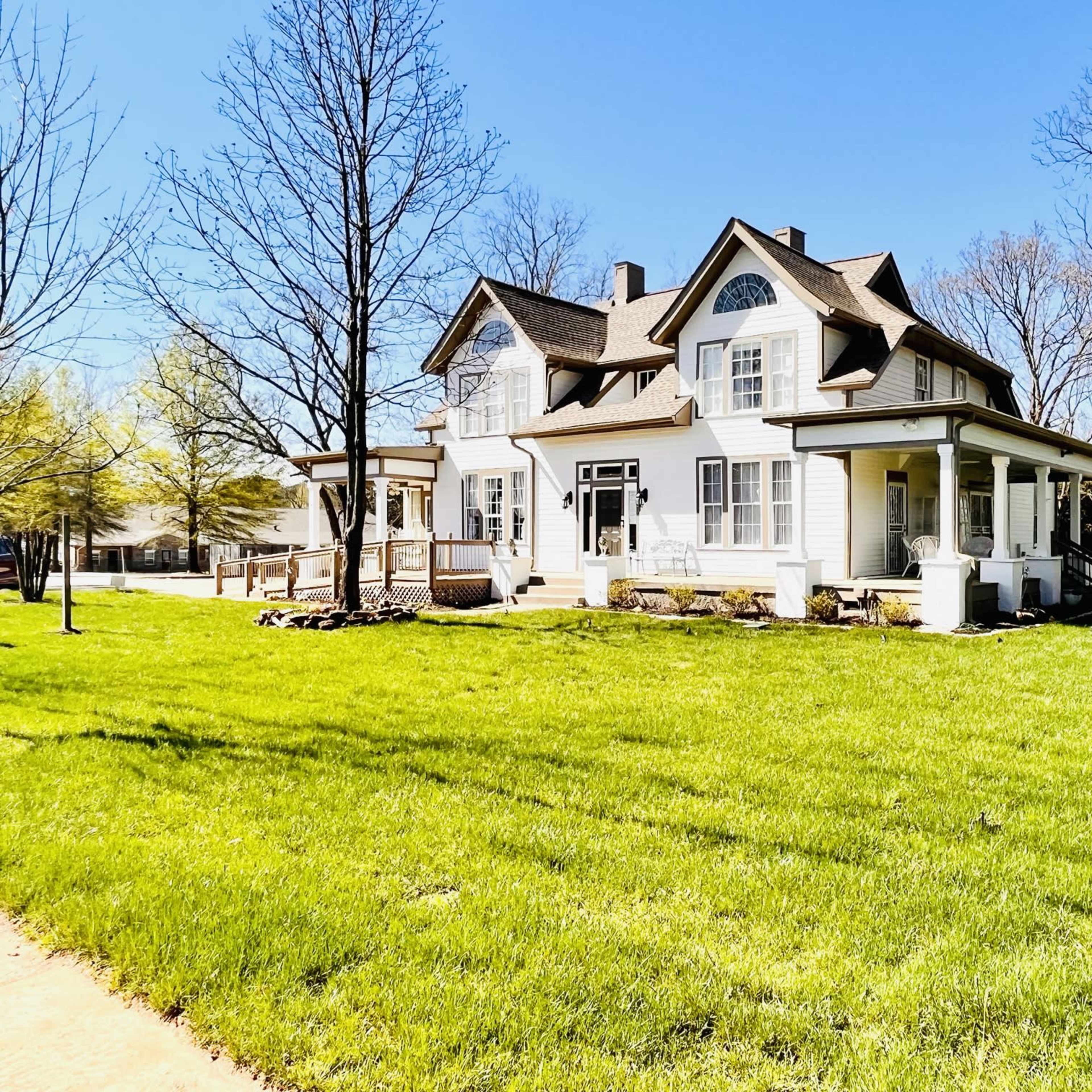 A large, two-story white house with a wraparound porch is situated on a grassy lawn surrounded by trees.