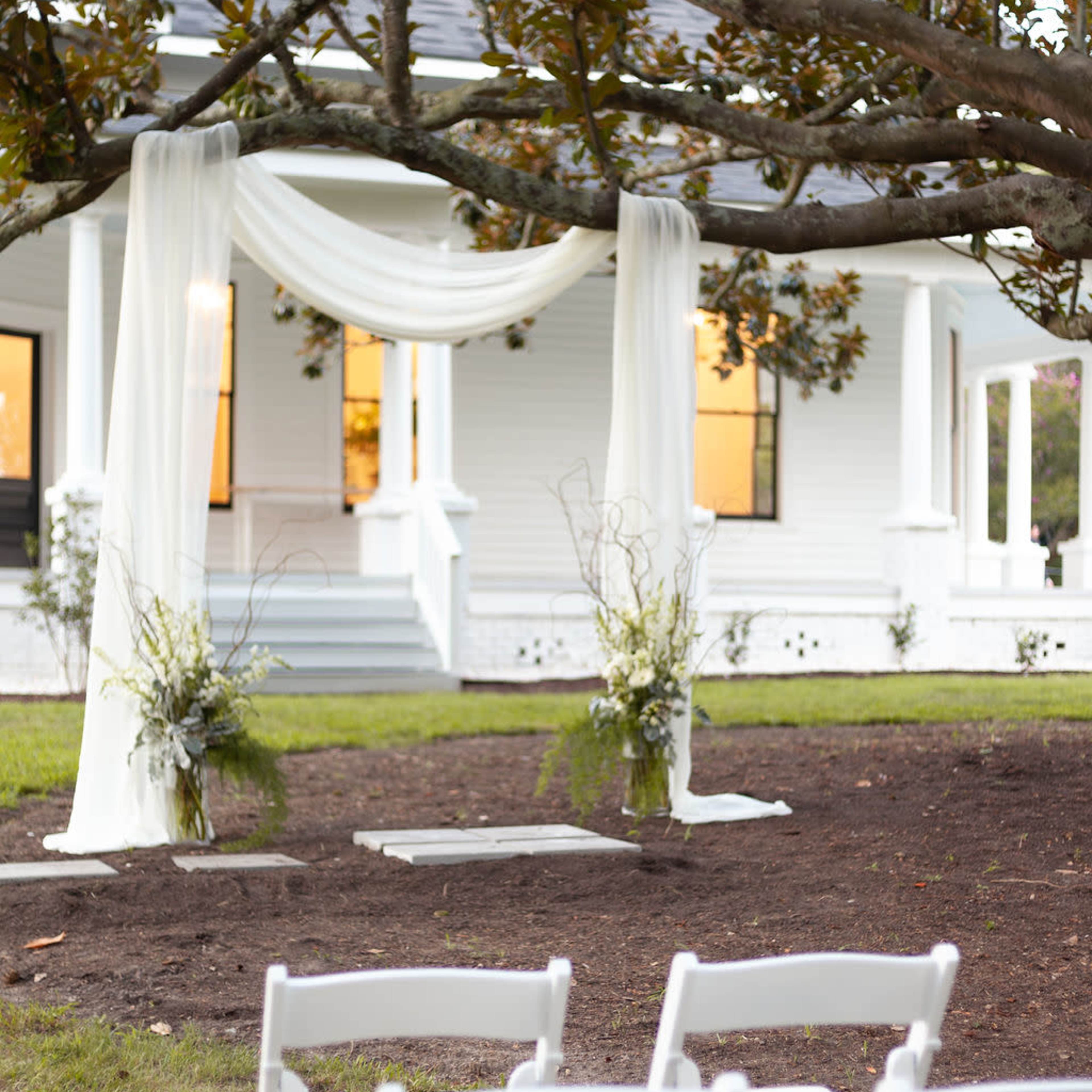 An outdoor wedding setup features a decorative draped arch under a large tree, with white chairs arranged in front on a grassy area.