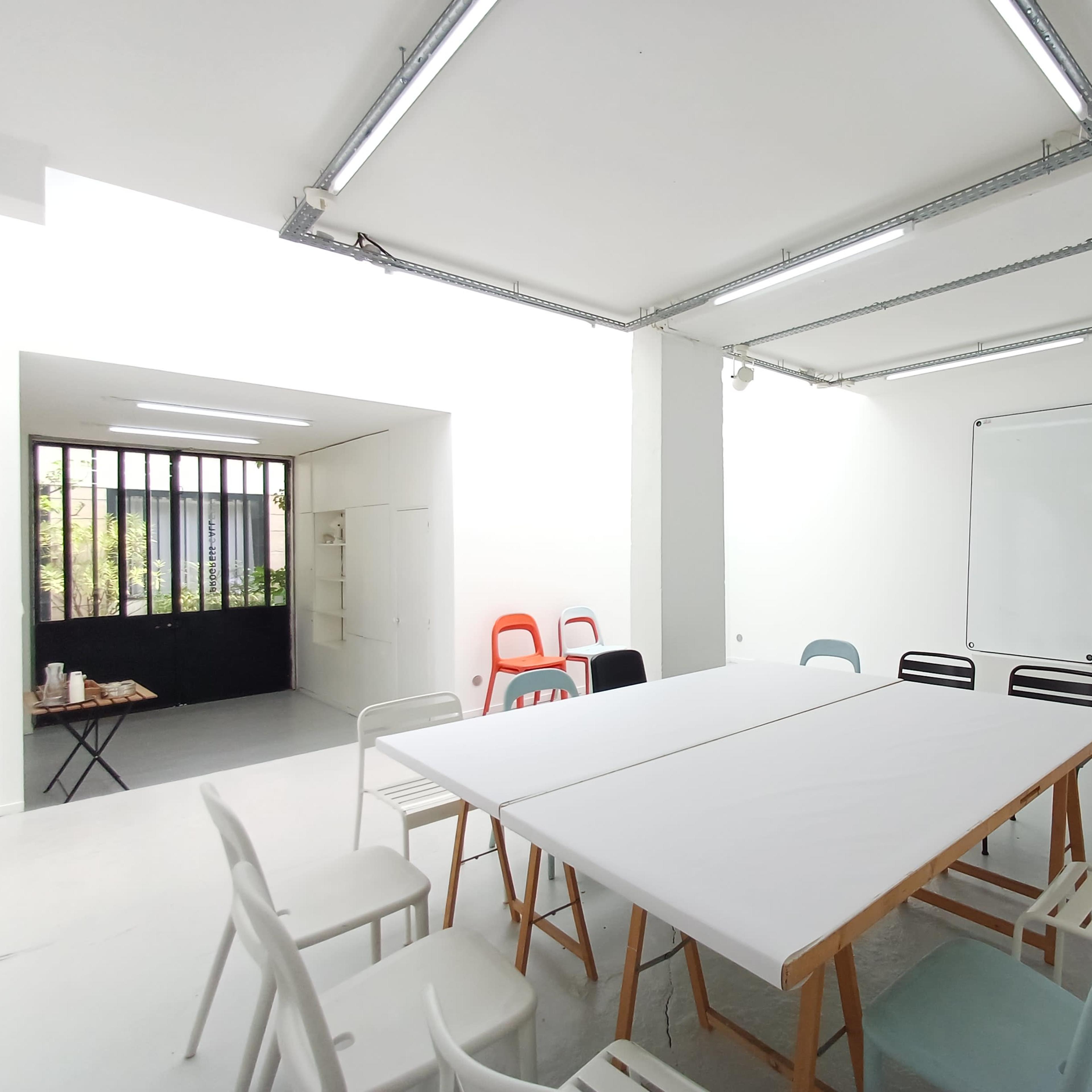 A brightly lit meeting room features a large table surrounded by various colored chairs, with a whiteboard on the wall and an open doorway leading to an outdoor space.