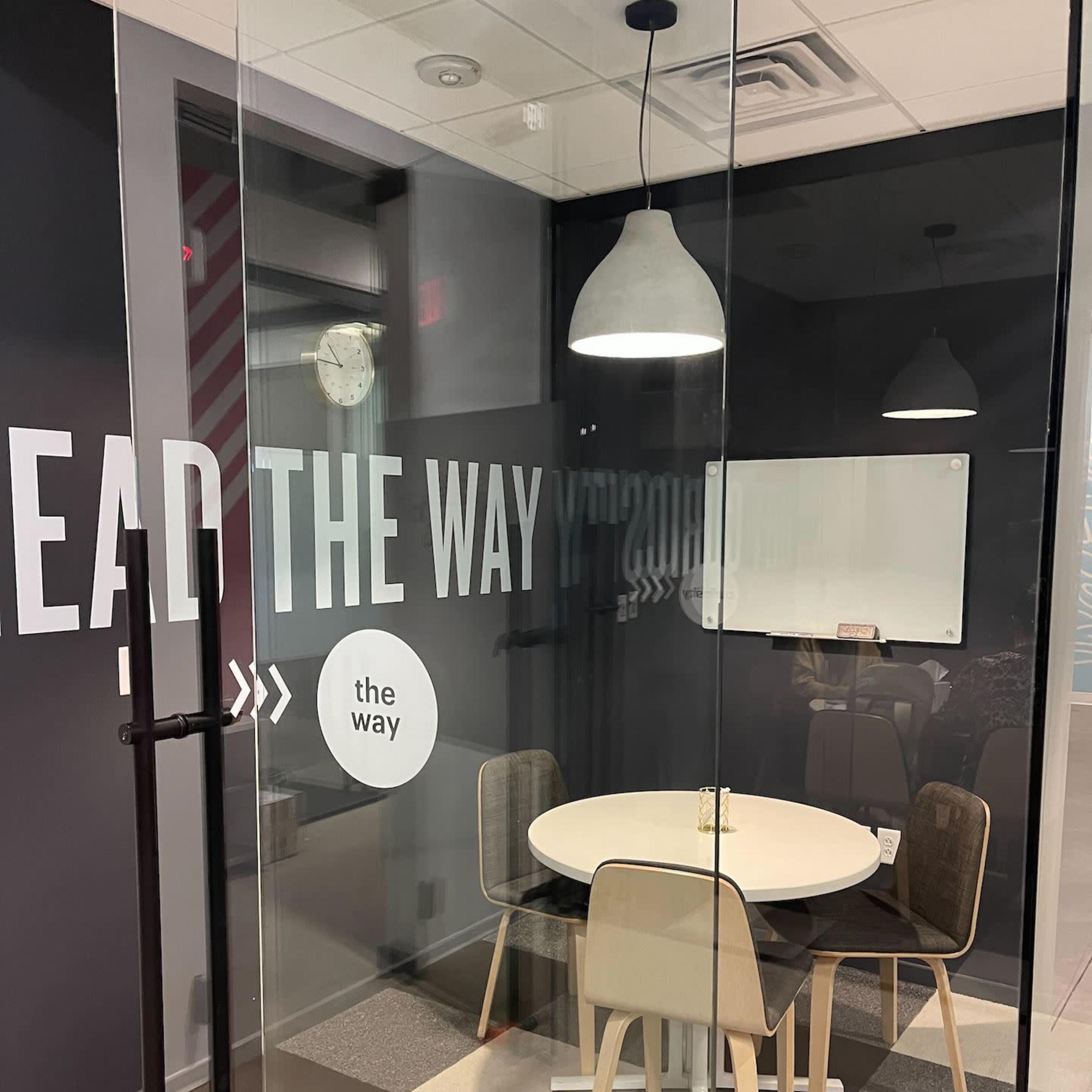 A glass-walled meeting room features a round table surrounded by four chairs, with a whiteboard and wall graphics.