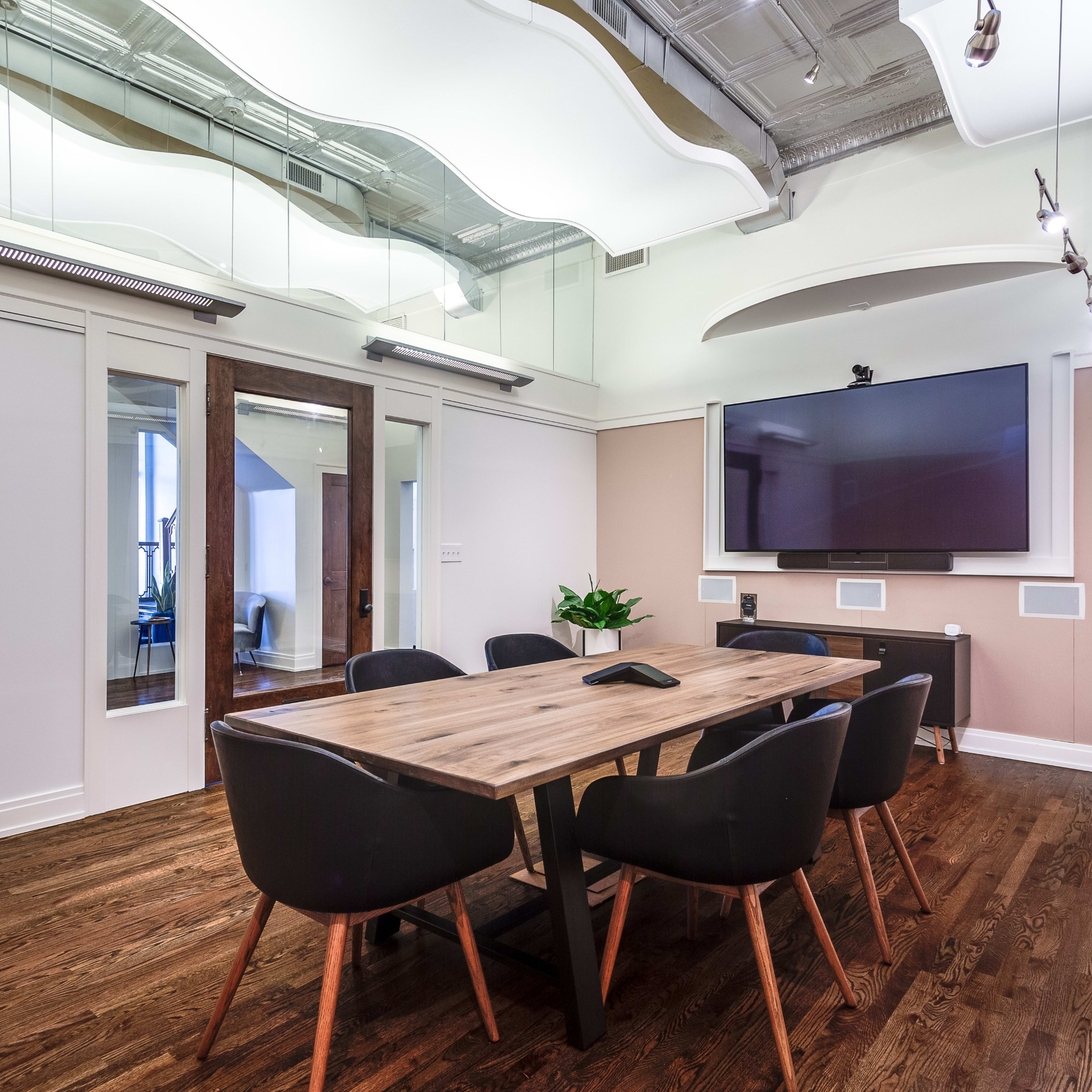 A modern conference room features a wooden table surrounded by black chairs, with a wall-mounted television and large windows allowing natural light.