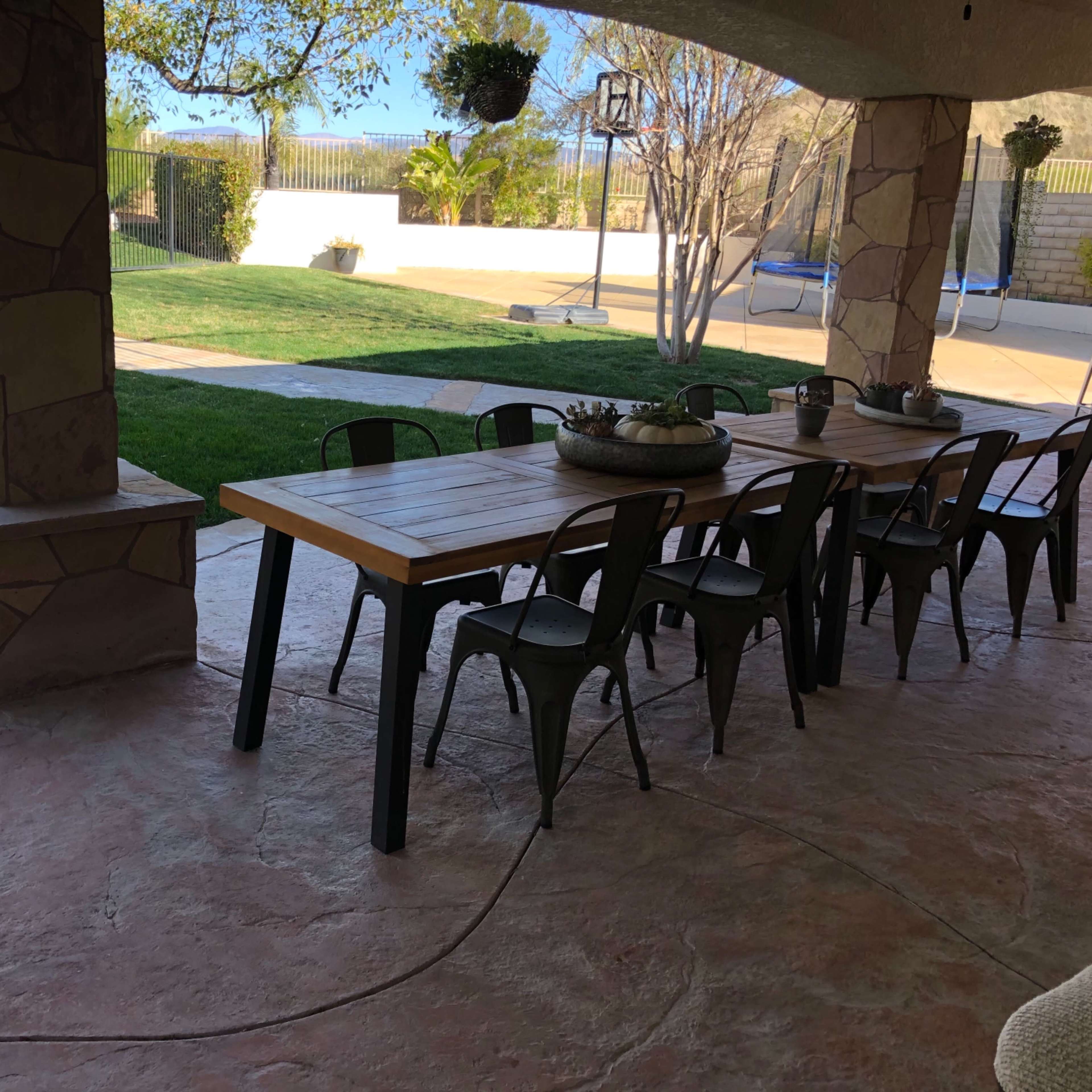 A long wooden dining table with black metal chairs is set up under a shaded patio near a green lawn.