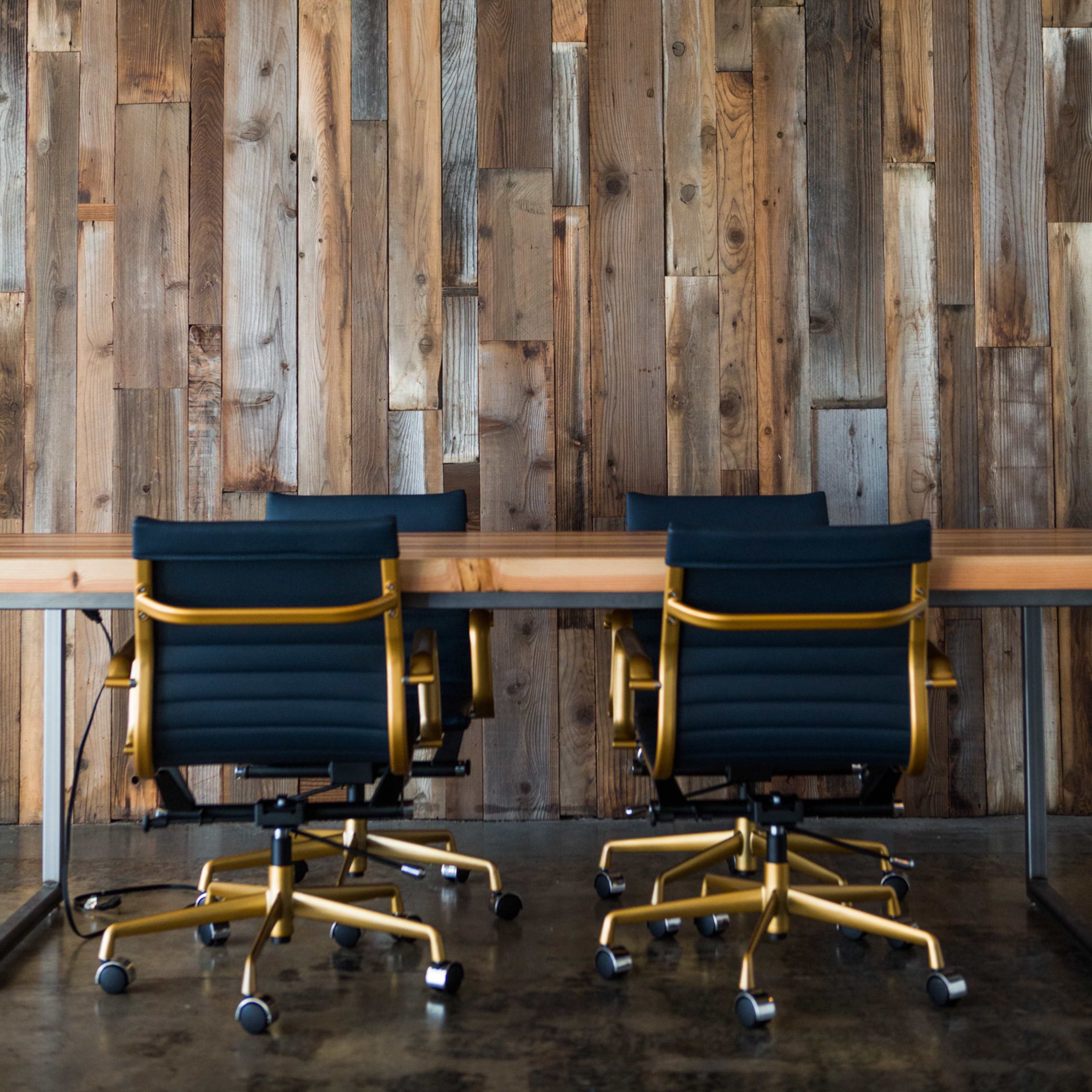 Two black office chairs with gold frames are positioned at a long wooden table against a backdrop of vertical wooden planks.