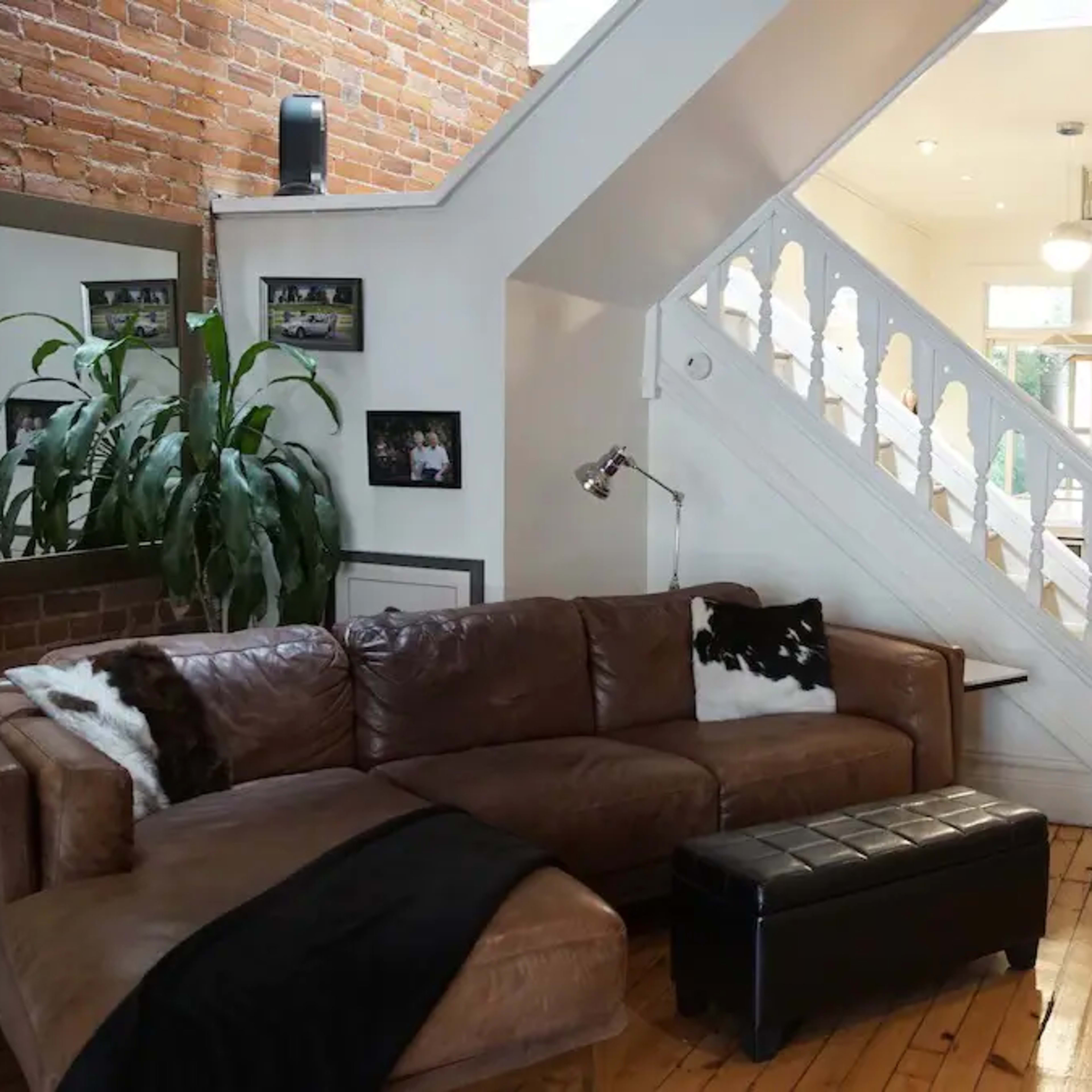 A well-lit living room with a brown leather couch, a black ottoman, and a staircase leading to an upper level, with a view of a hallway and an orange door.