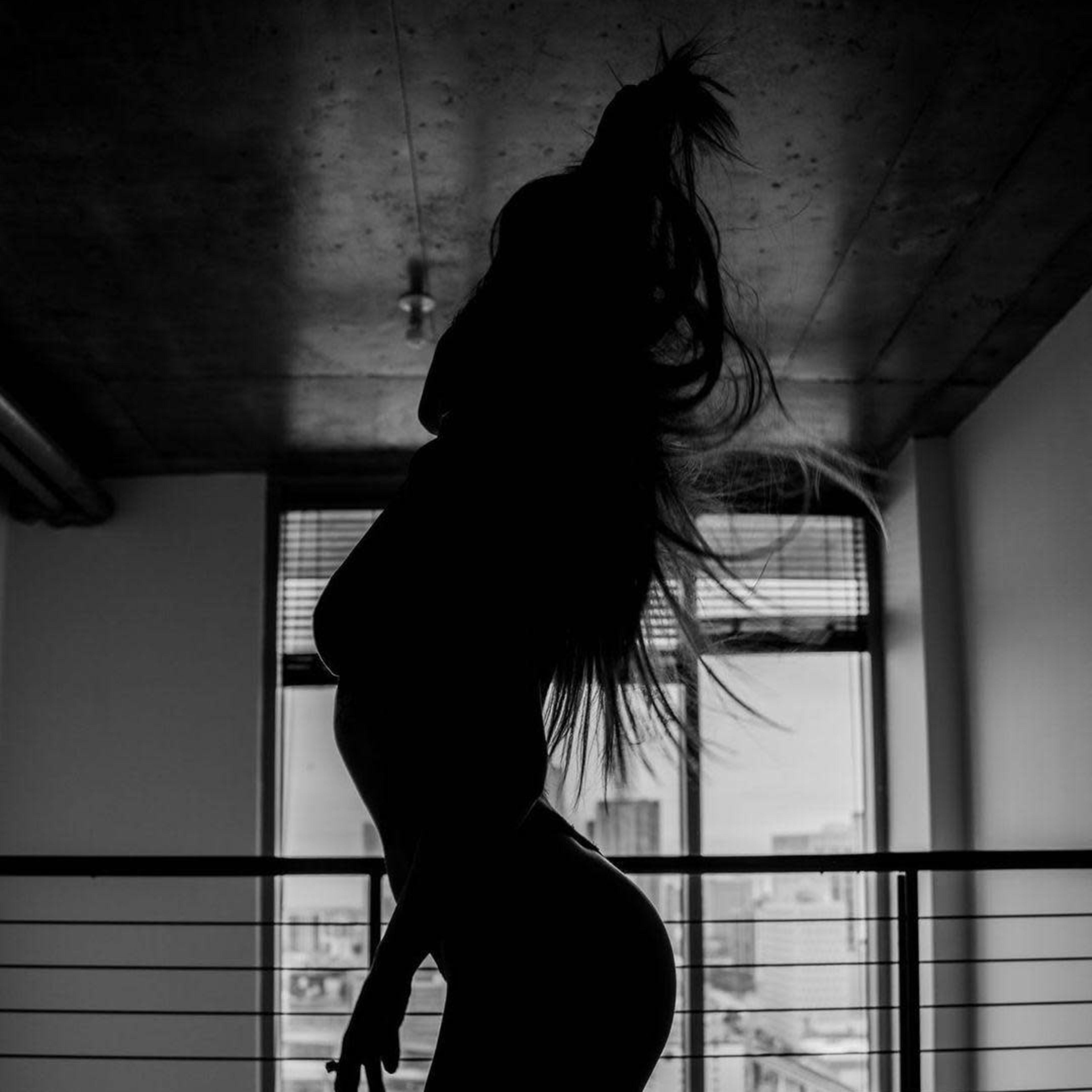A silhouetted figure kneels on a stool in front of large windows with urban scenery visible outside.