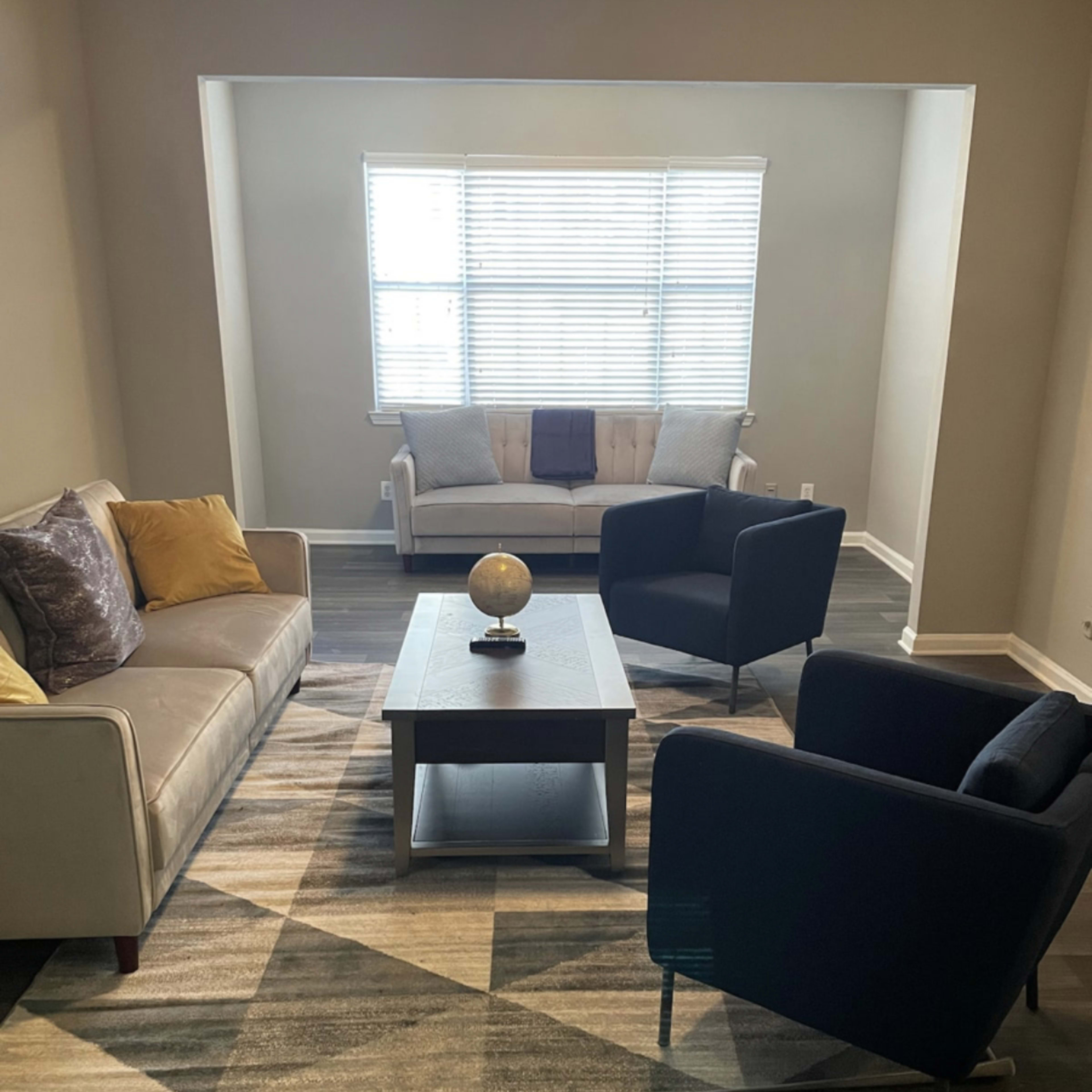 A living room features a gray sofa, two black chairs, a coffee table, and a patterned rug beneath a window with blinds.