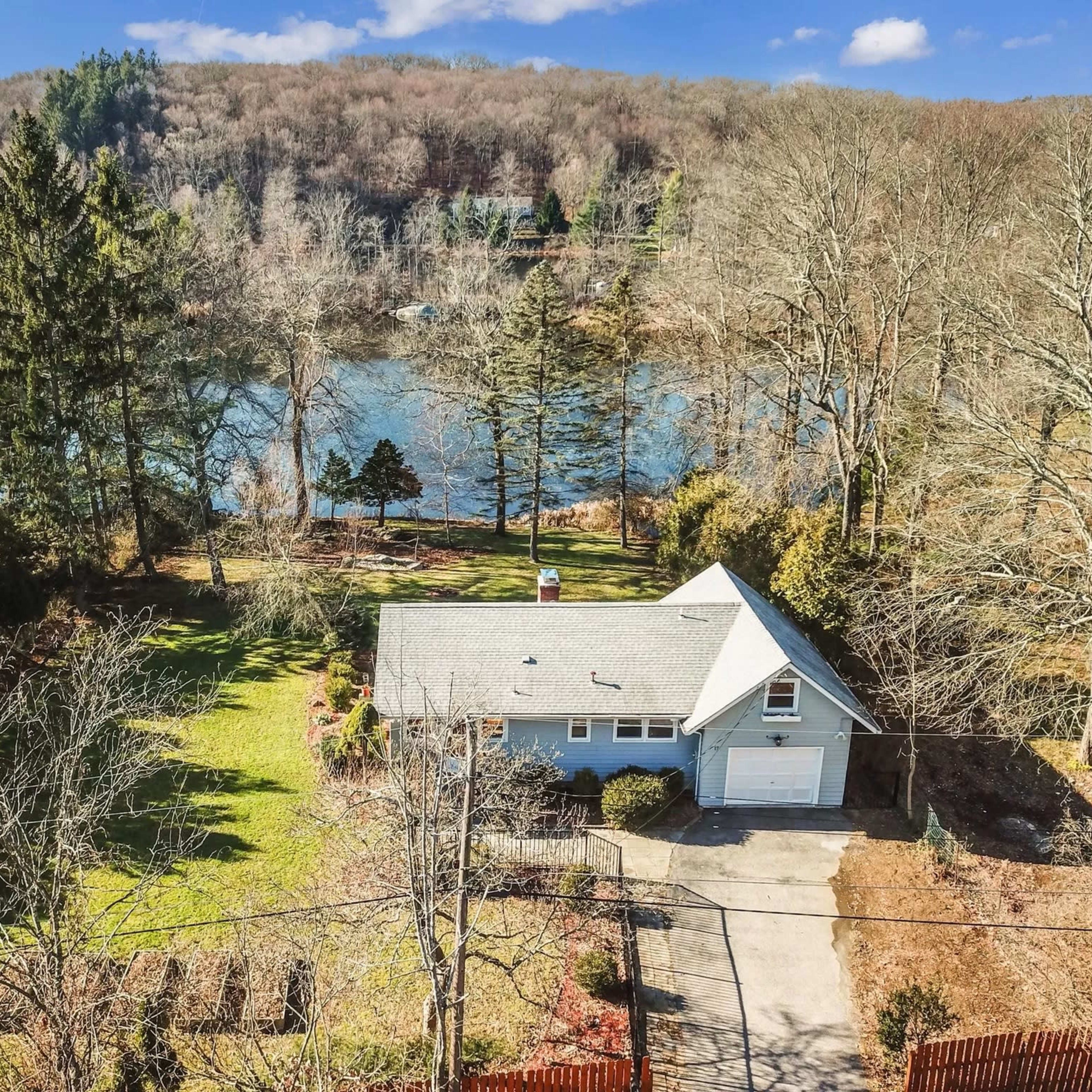 A single-story blue house with a garage is situated on a spacious lot near a calm body of water, surrounded by bare trees and rolling hills in the background.