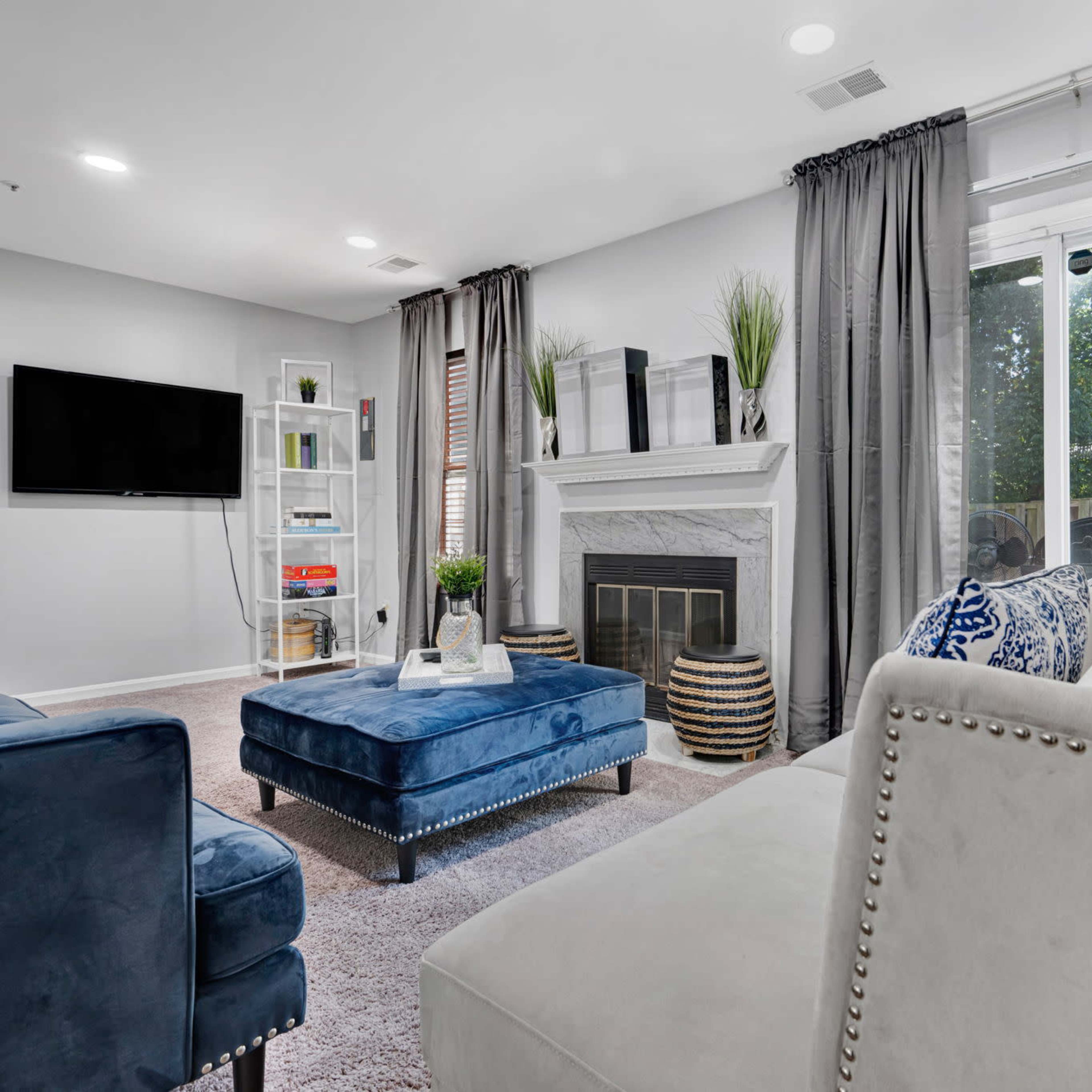 A modern living room features a white sofa, a blue ottoman, a fireplace, and floor-to-ceiling curtains, with a television mounted on the wall.