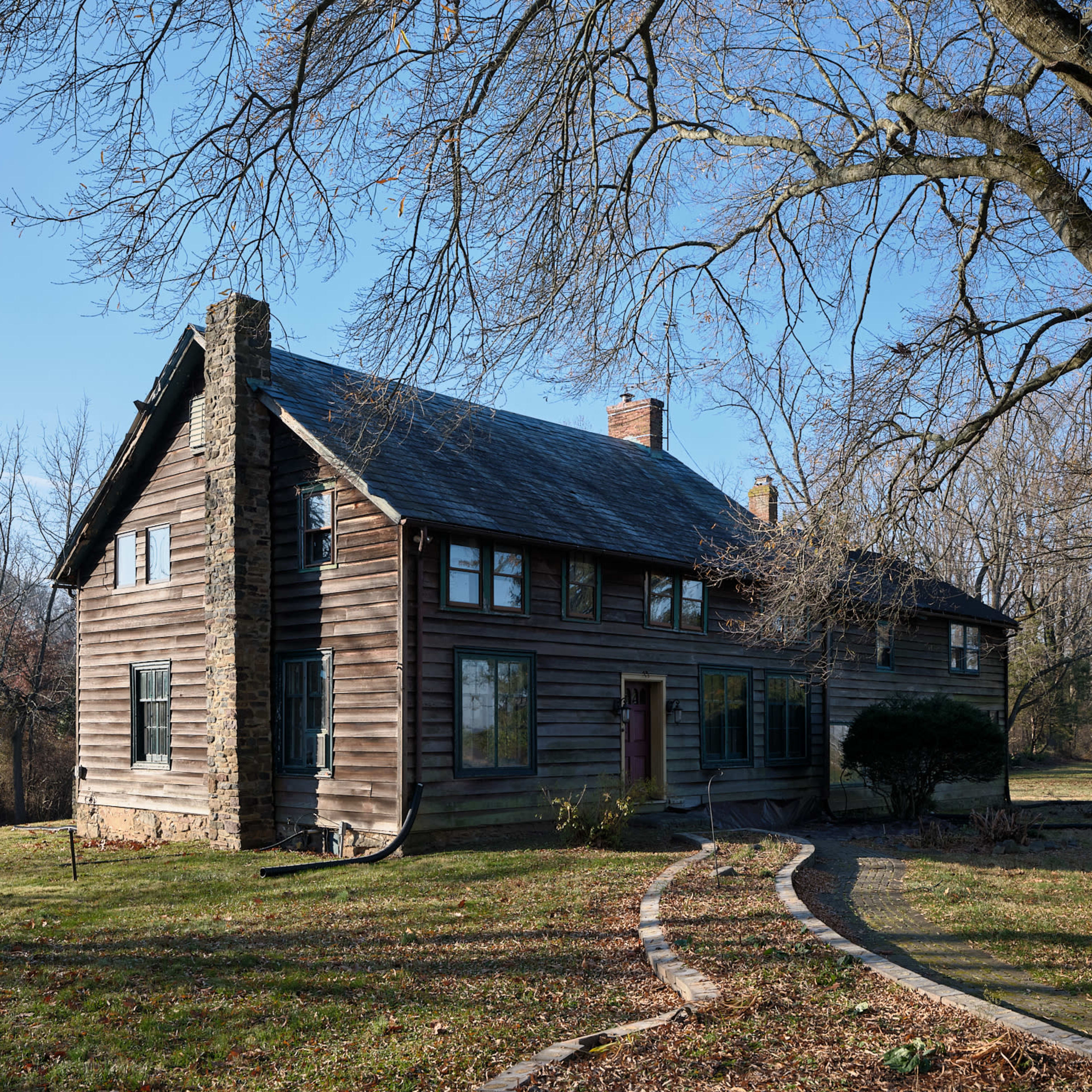 A two-story, wooden house with a stone chimney, set on a grassy plot with a winding stone pathway and bare trees in the background.