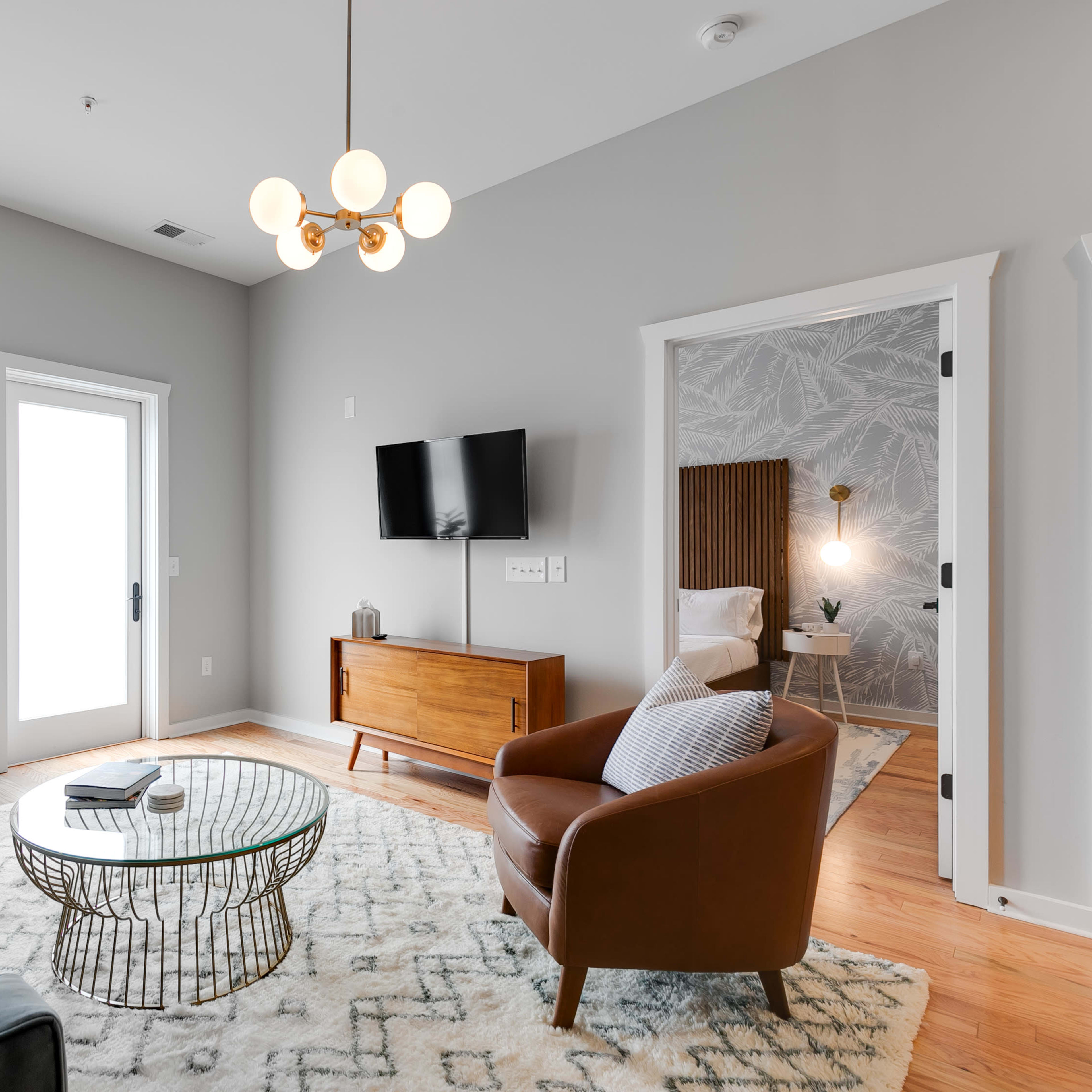 The living room features a blue sofa, a brown leather armchair, a round coffee table, and a television mounted on the wall, with green curtains and a plant by the window.