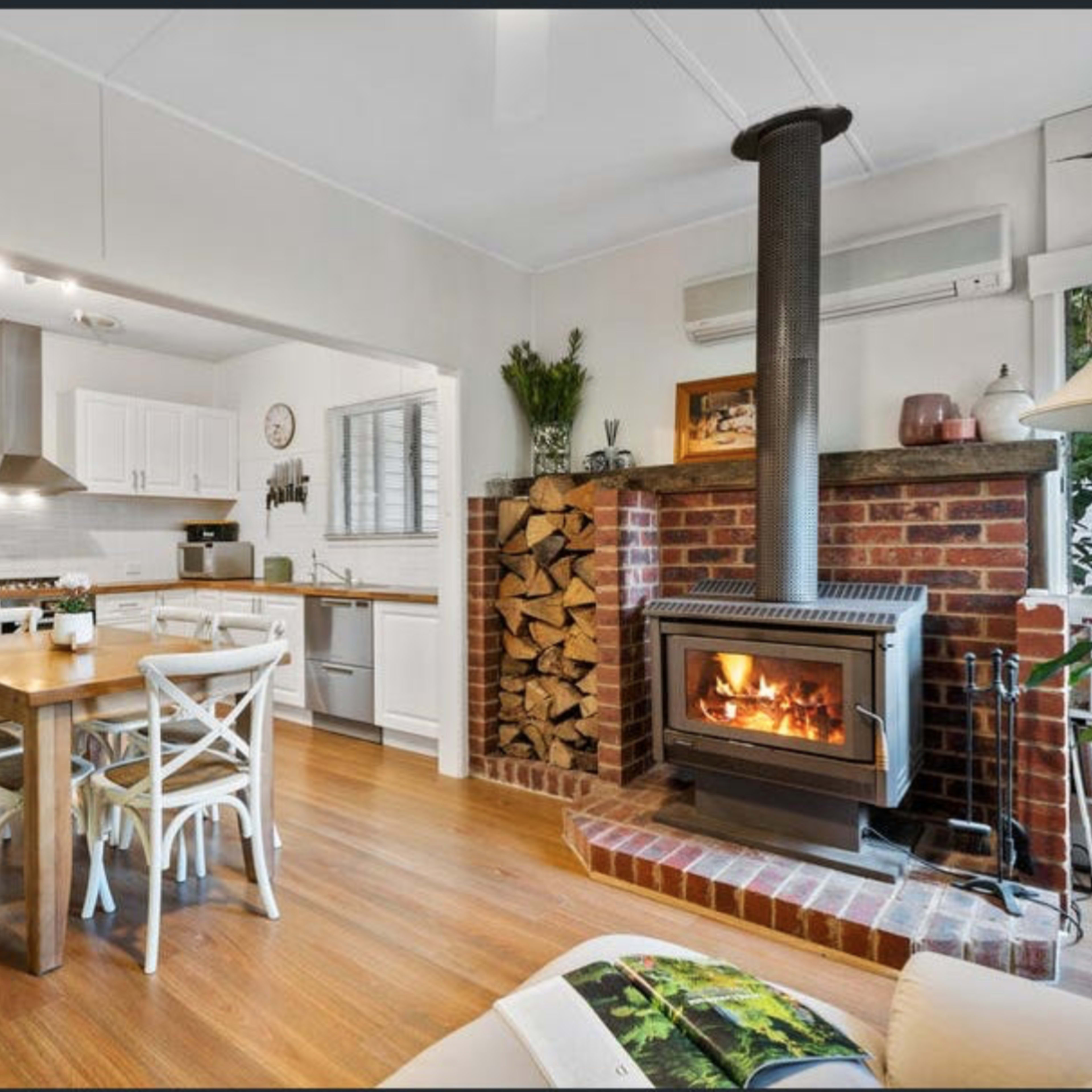 A cozy kitchen and dining area featuring a wooden table with chairs, a brick fireplace with a wood stack beside it, and modern appliances in a bright, open layout.