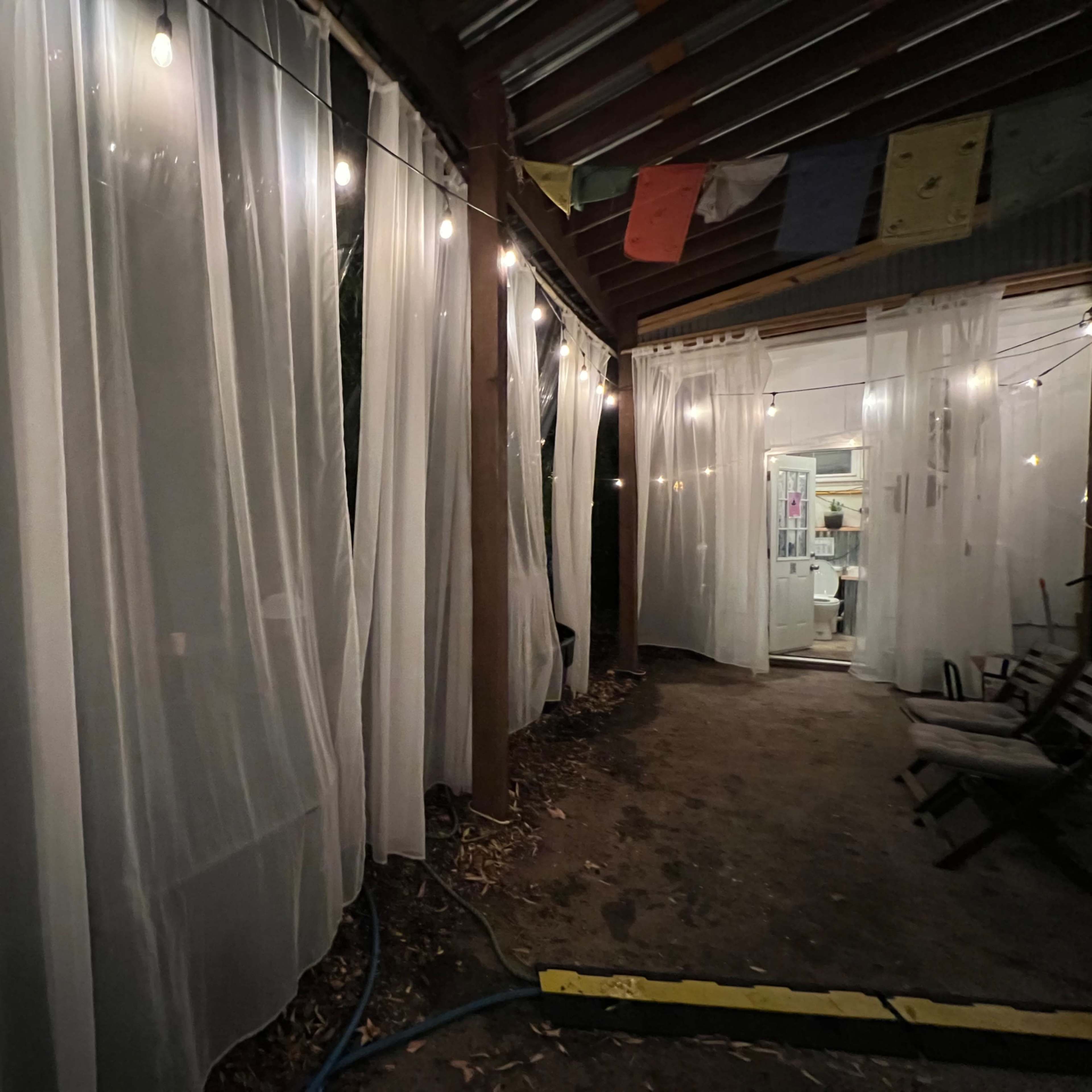 A dimly lit outdoor space with sheer white curtains and string lights hanging from a wooden structure.