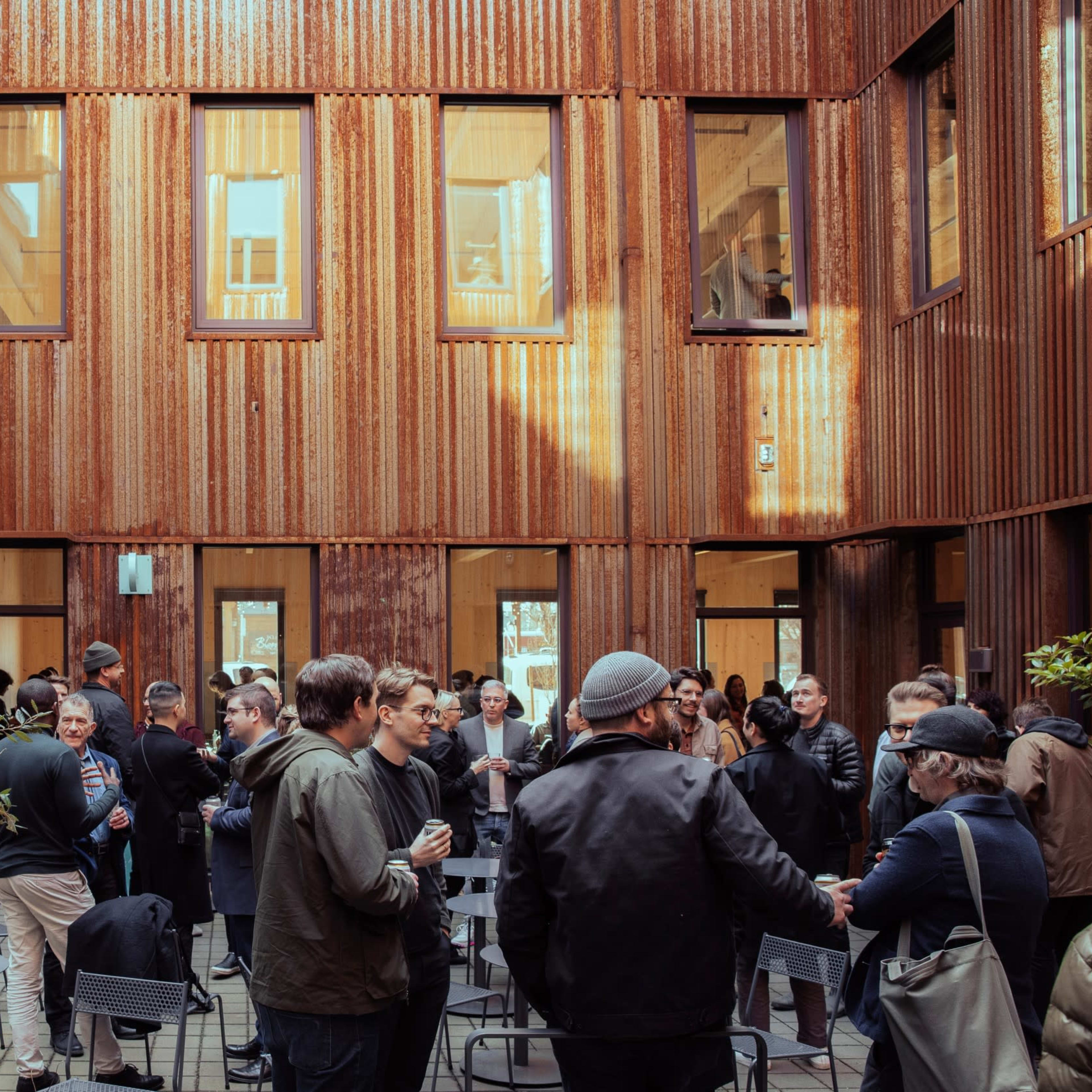 A group of people gathers in a courtyard surrounded by a modern building with wooden siding and multiple windows.