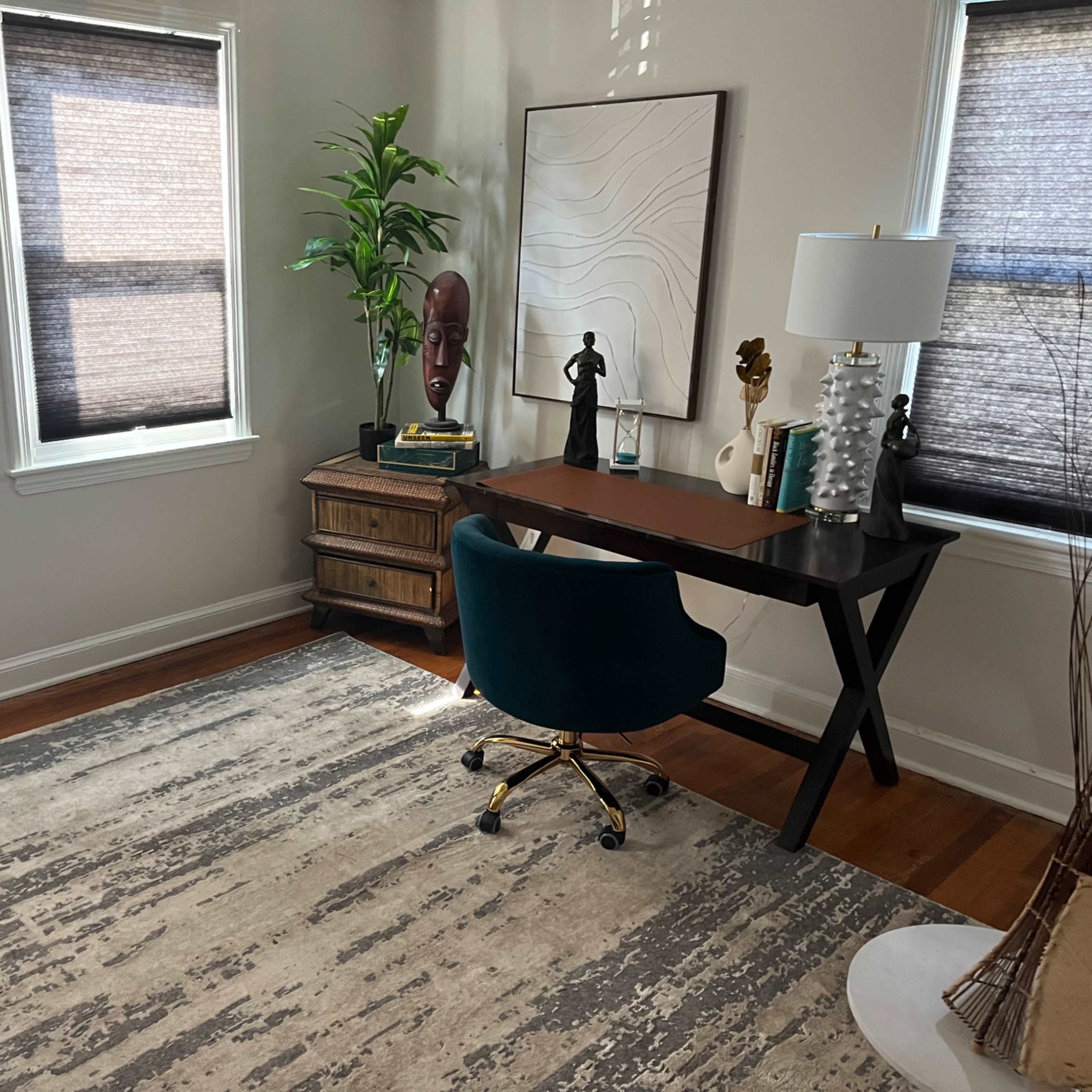 A home office features a dark wooden desk with a turquoise chair, a decorative plant, and art displayed on the walls, along with a light-colored area rug on the wooden floor.