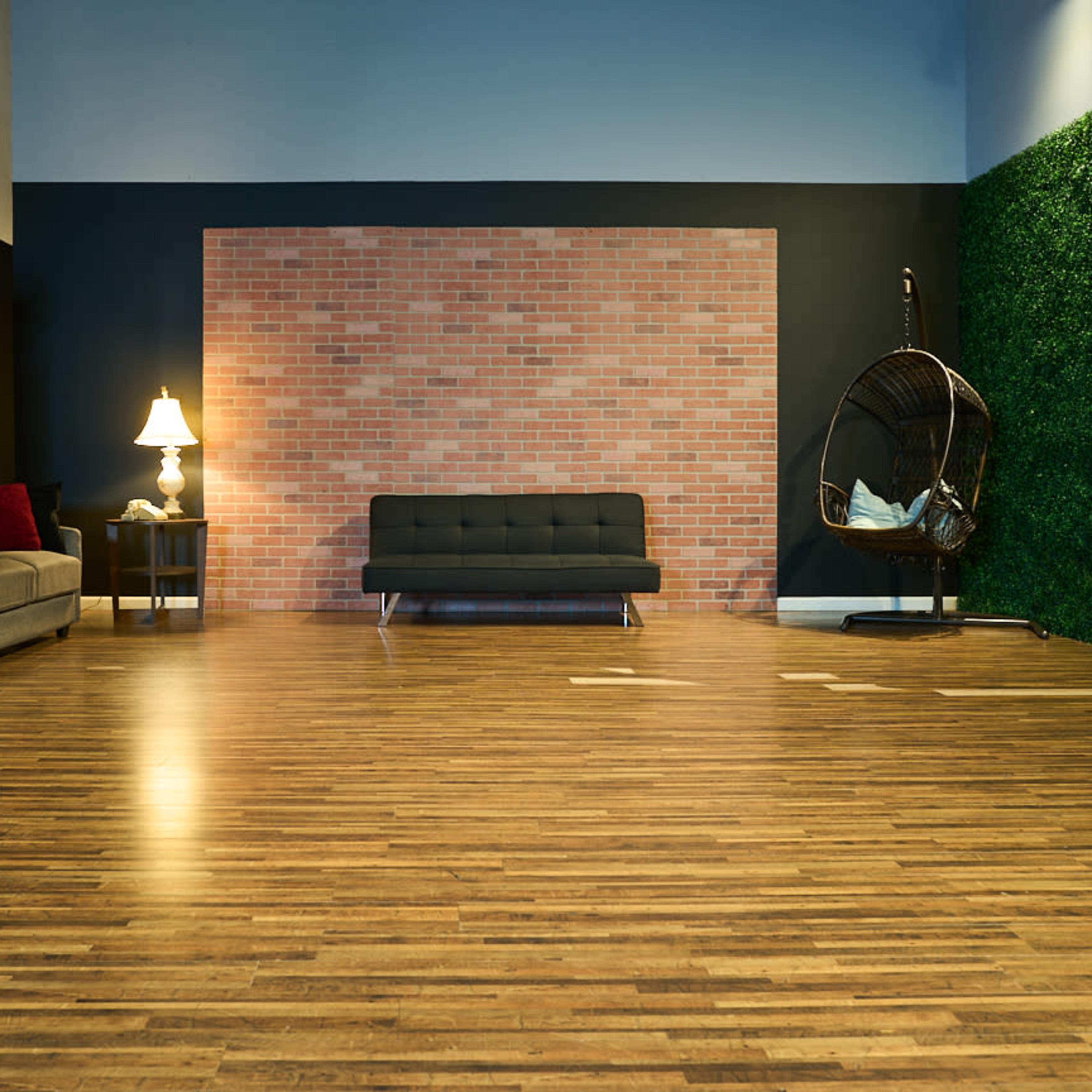 A spacious interior featuring a wooden floor, a gray sofa, a black couch, a hanging chair, a lamp, and a wall with a brick pattern and artificial greenery.