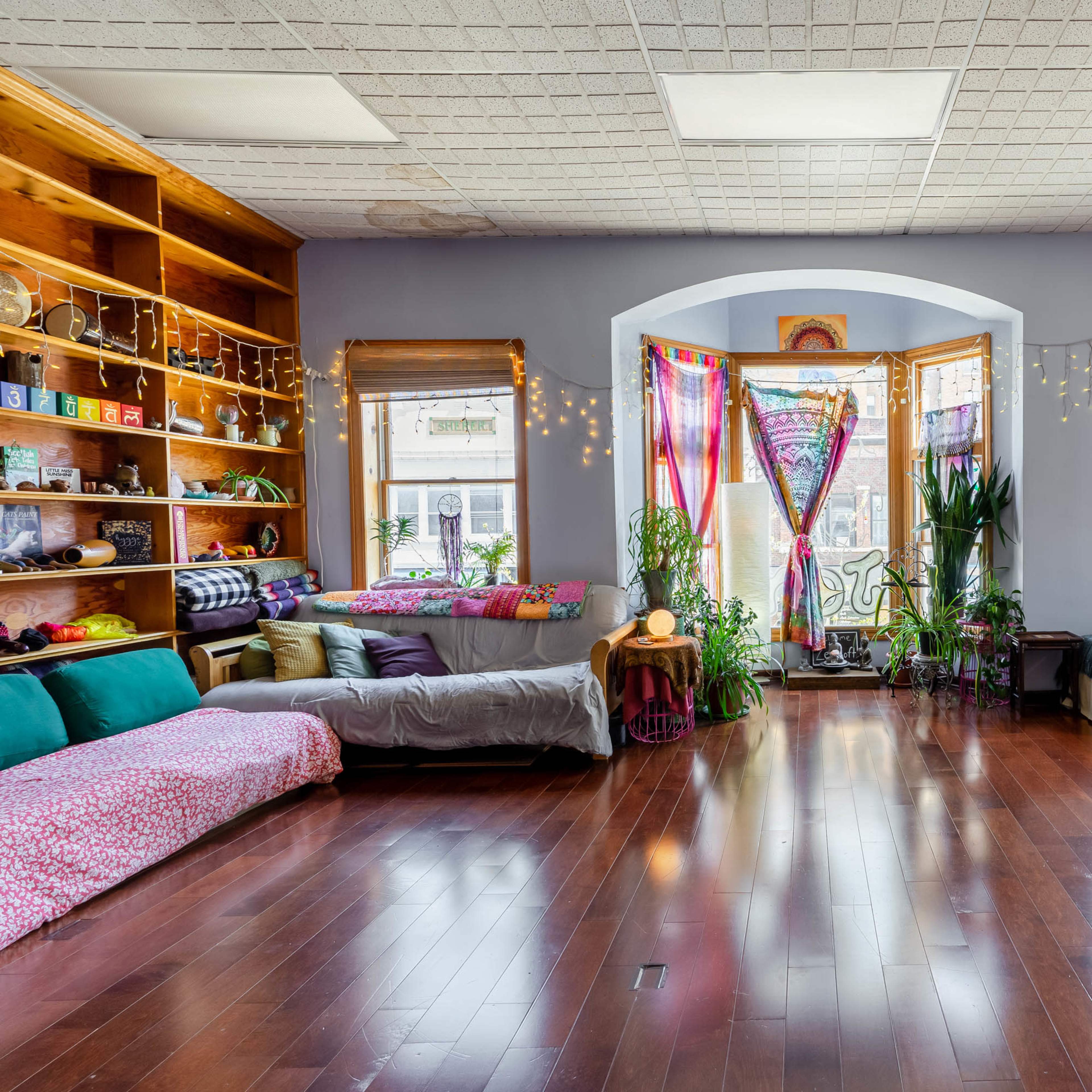 A spacious room with wooden shelves filled with books and colorful cushions arranged around large windows adorned with plants and fabric drapes.