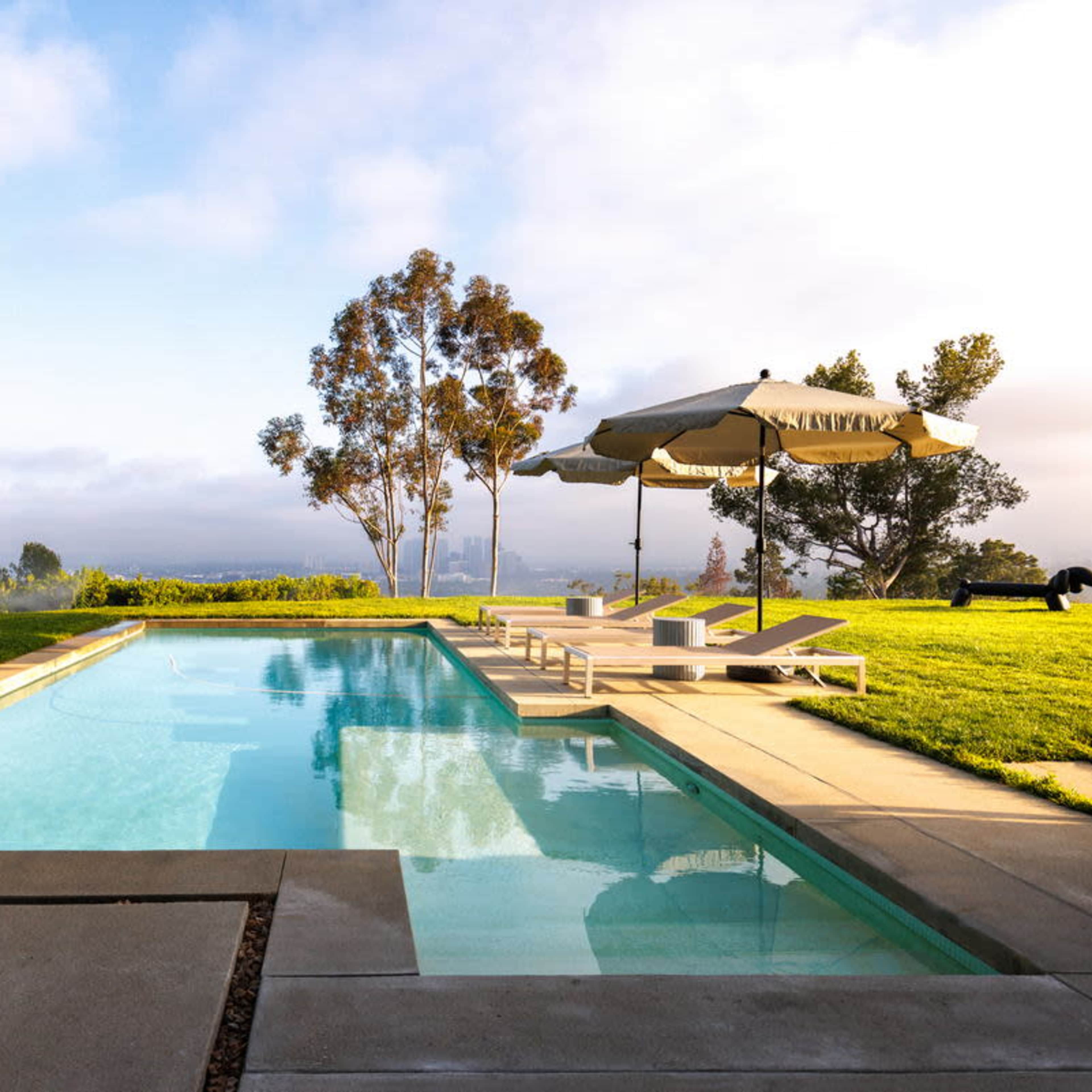 A modern swimming pool is surrounded by lounge chairs and umbrellas, set against a backdrop of greenery and a cloudy sky.