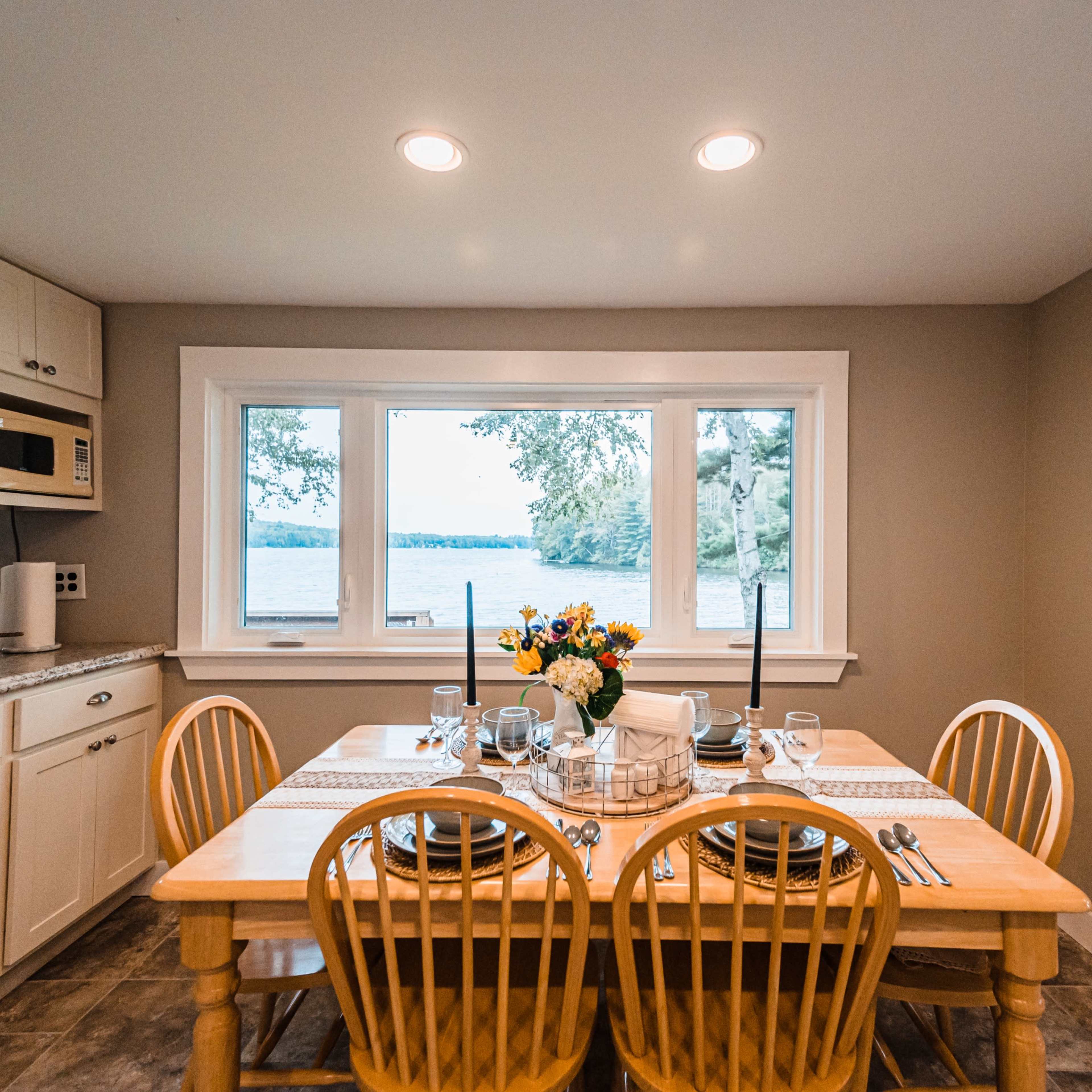 A dining area features a wooden table set for a meal, with a bouquet of flowers, and large windows overlooking a lake.