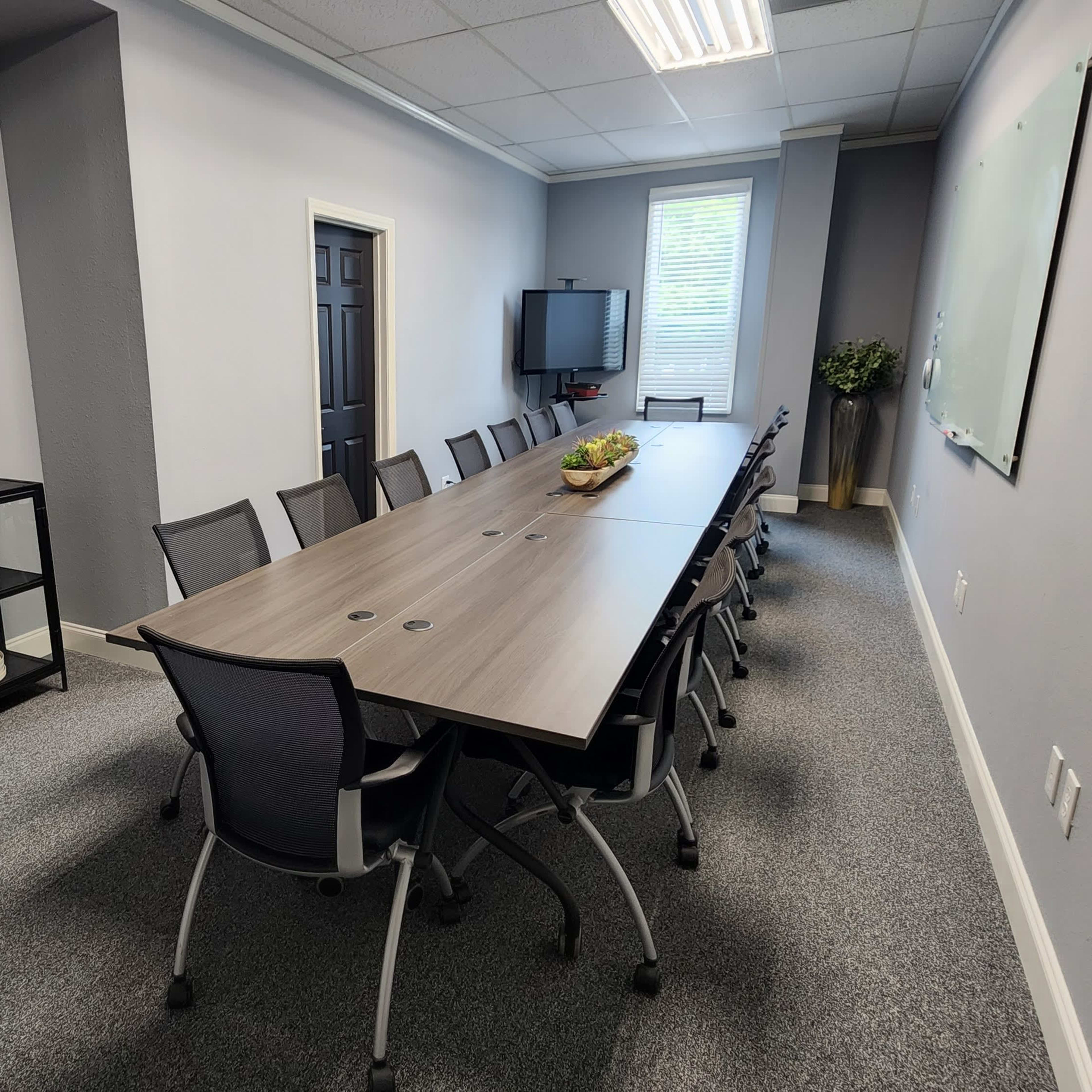 A conference room features a long table surrounded by chairs, with artwork on the wall and a television in the corner.