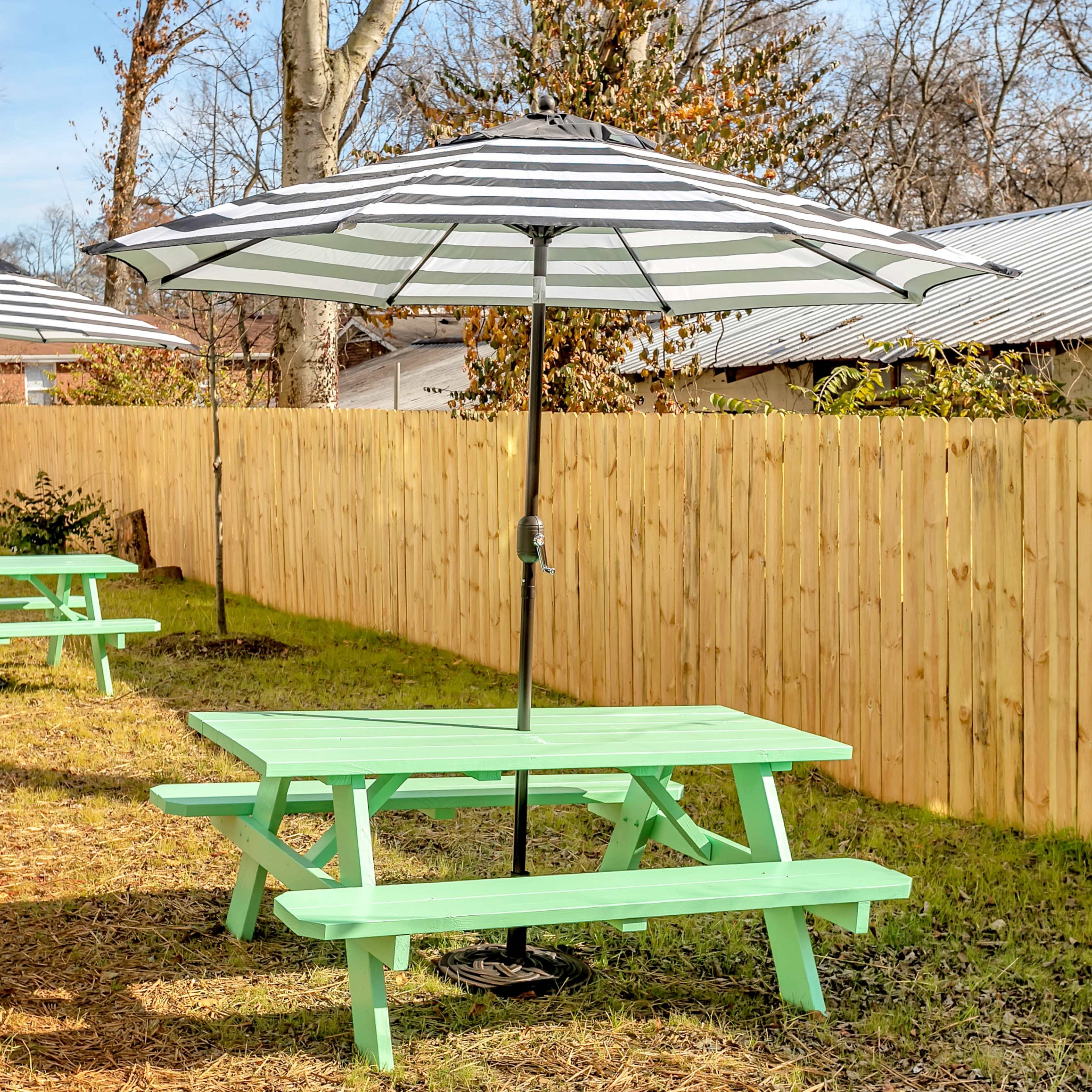 Two green picnic tables with striped umbrellas set in a grassy area next to a wooden fence.