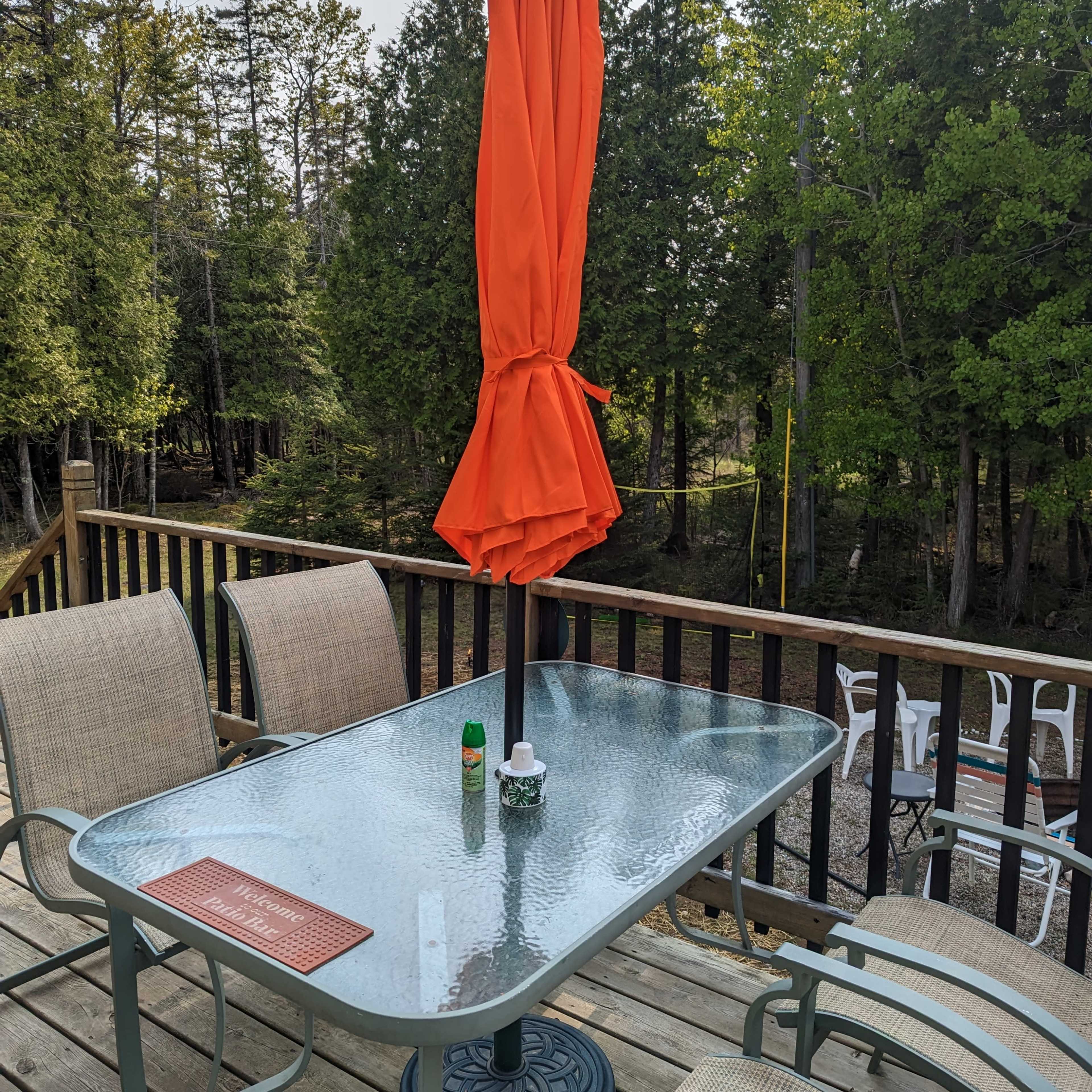 A glass-top table with an orange umbrella and four chairs is set on a deck overlooking a wooded area.