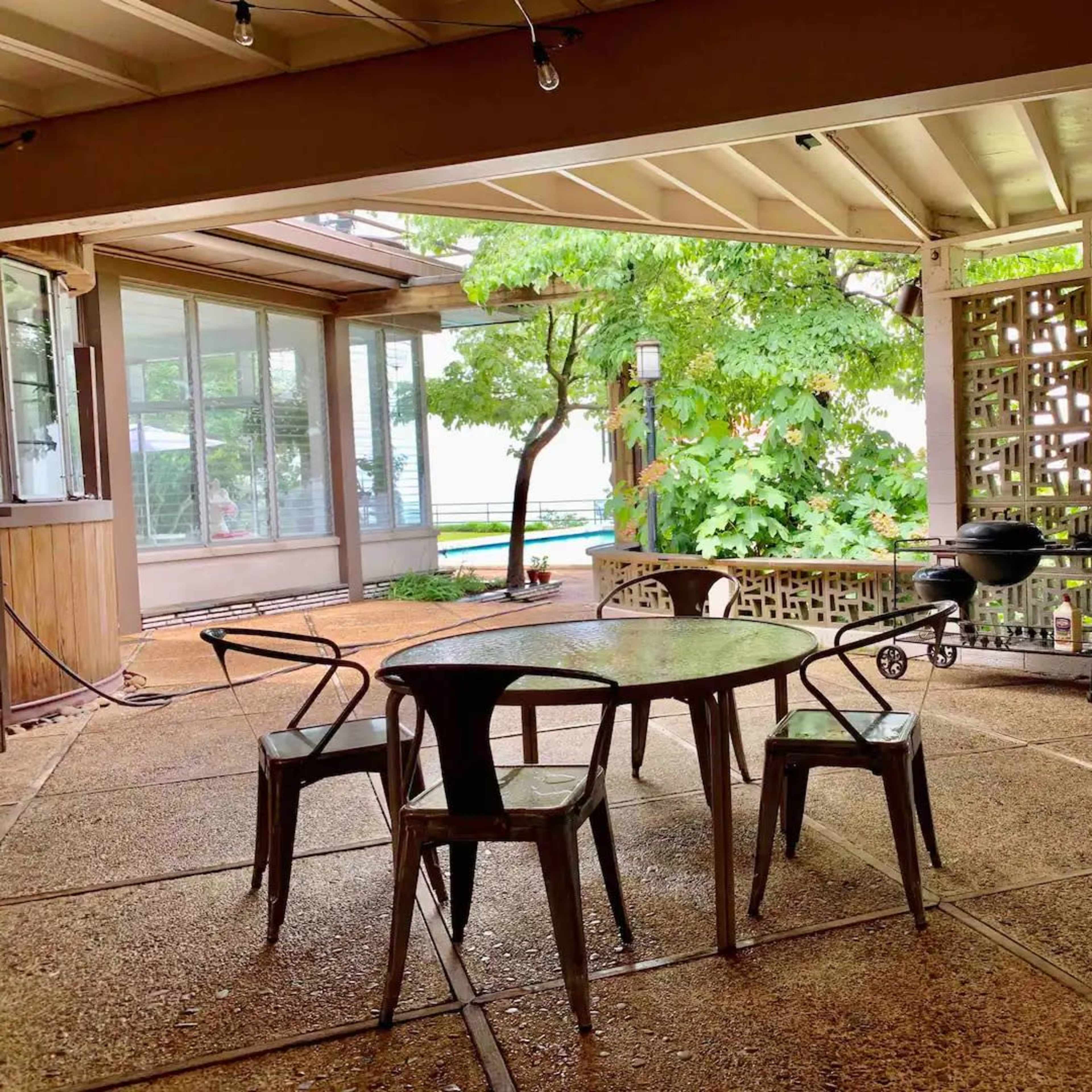A covered outdoor patio features a round table with four metal chairs and a small grill, surrounded by greenery.