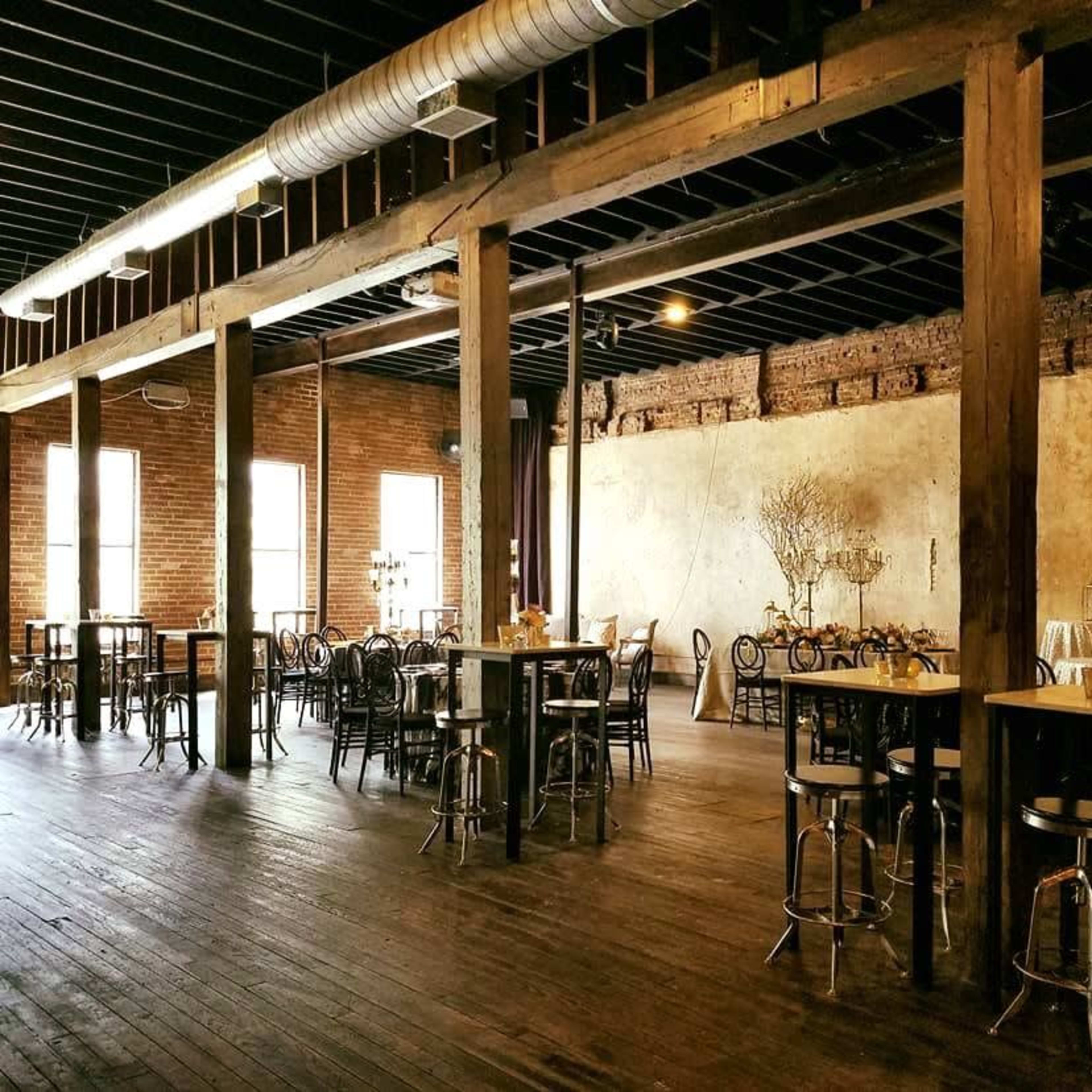 An empty industrial-style event space with wooden beams, brick walls, and several tables scattered throughout.