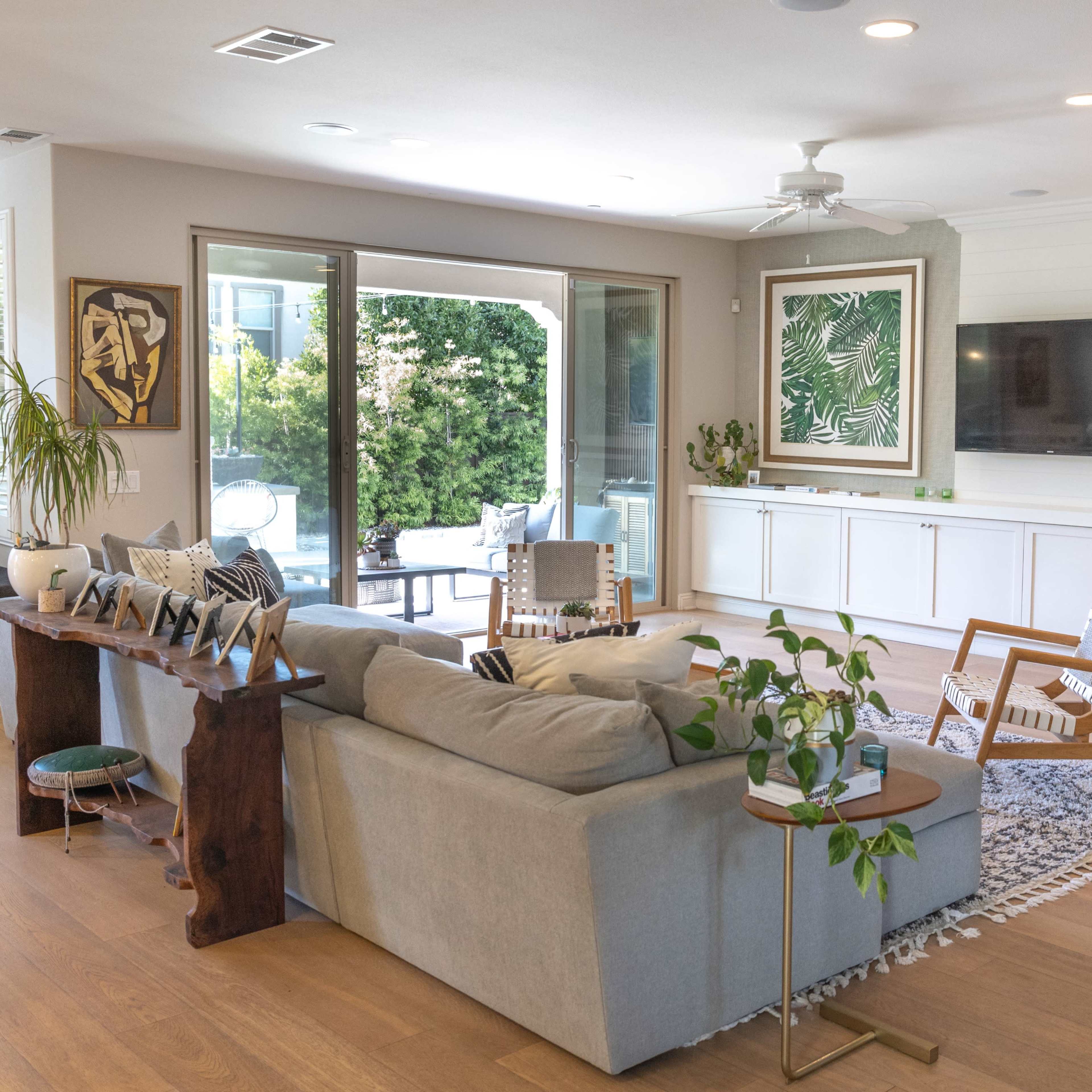 A modern living room with a gray sofa, wooden accent table, and sliding glass doors leading to an outdoor area, featuring large green-framed art pieces on the walls.
