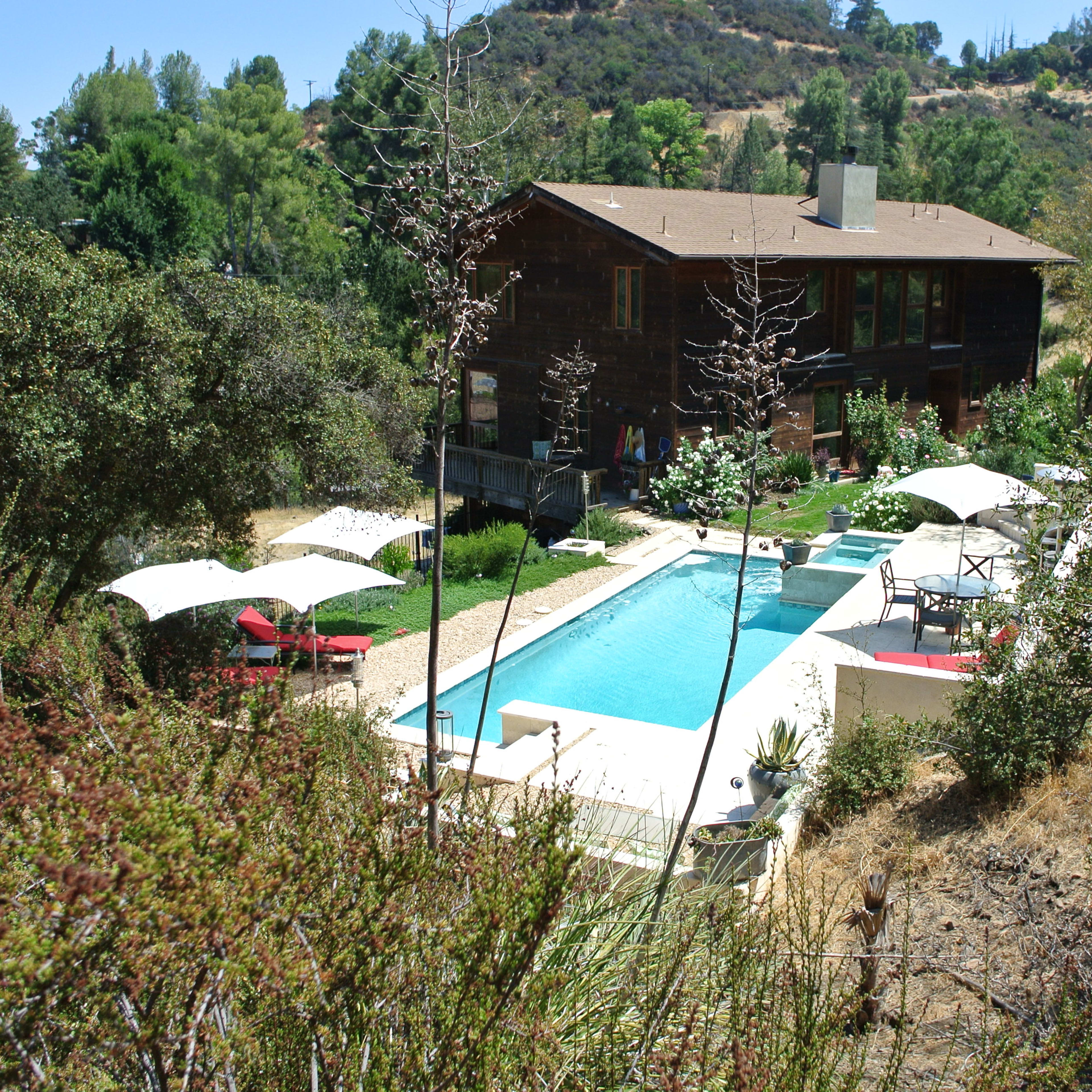 A modern house with large windows overlooks a rectangular swimming pool surrounded by lounge chairs and umbrellas, situated in a hilly landscape with greenery.
