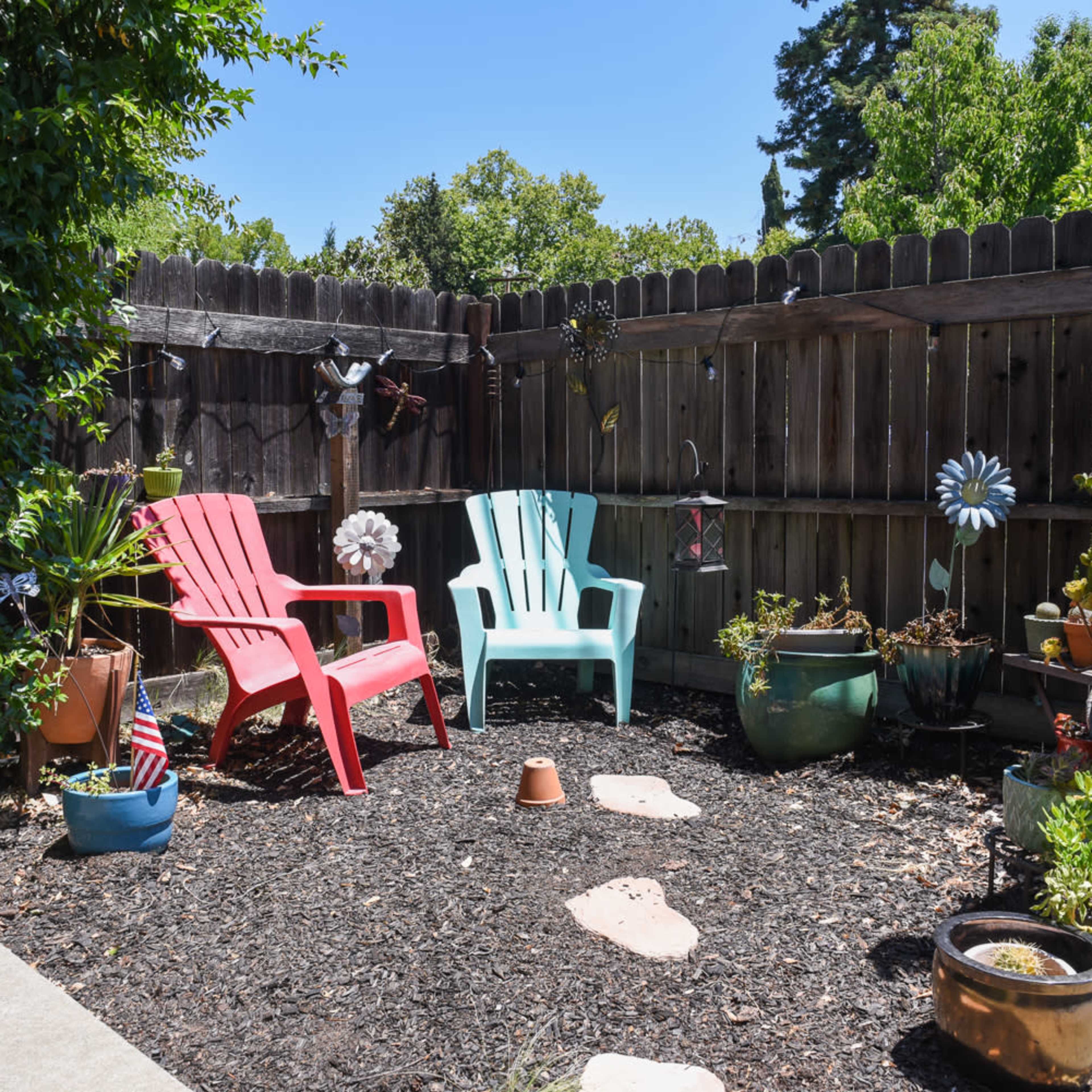 A small outdoor space with two plastic chairs, one red and one light blue, surrounded by potted plants and decorative items, enclosed by a wooden fence.