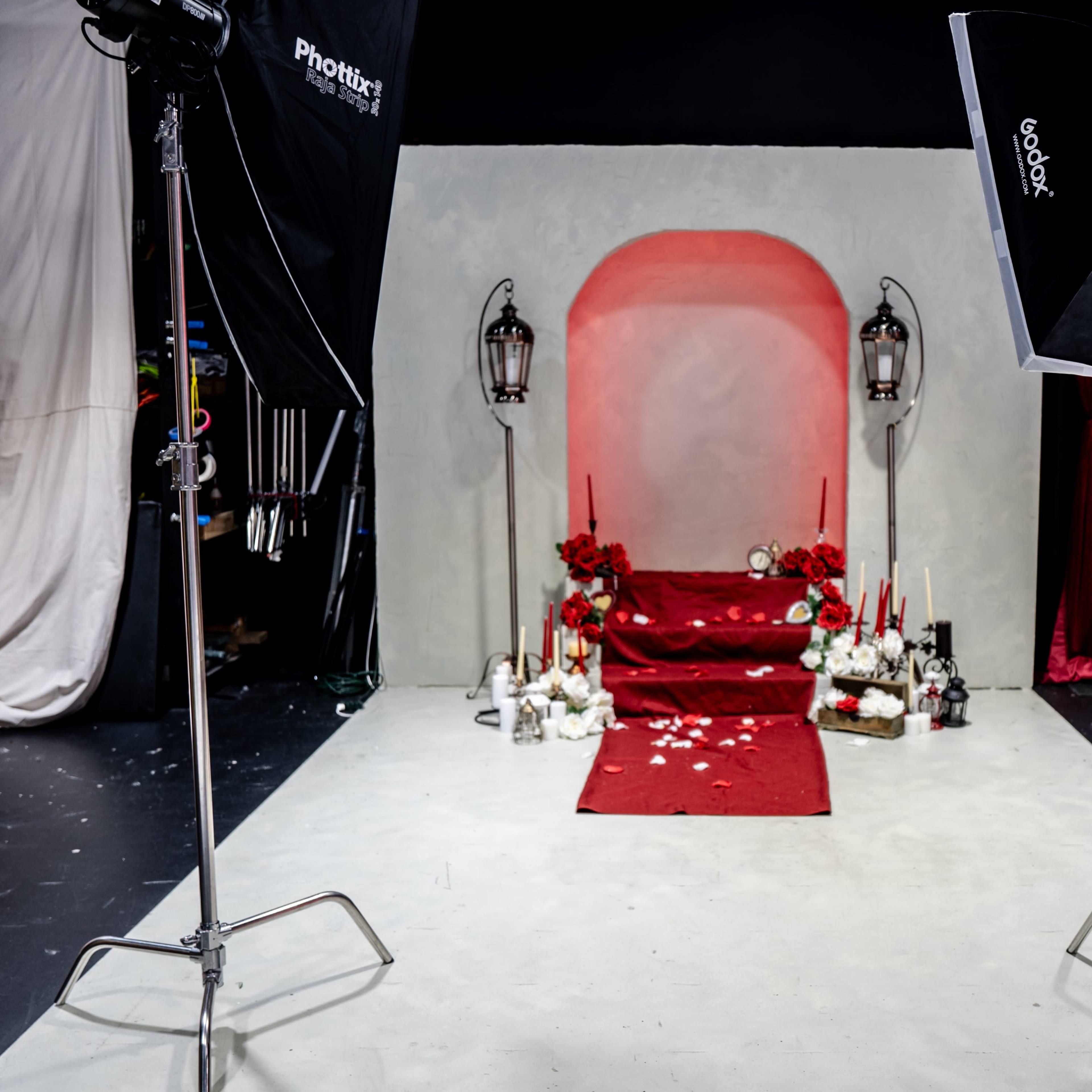 A photography studio setup with a red carpet leading to an arched backdrop adorned with lanterns, candles, and floral arrangements.