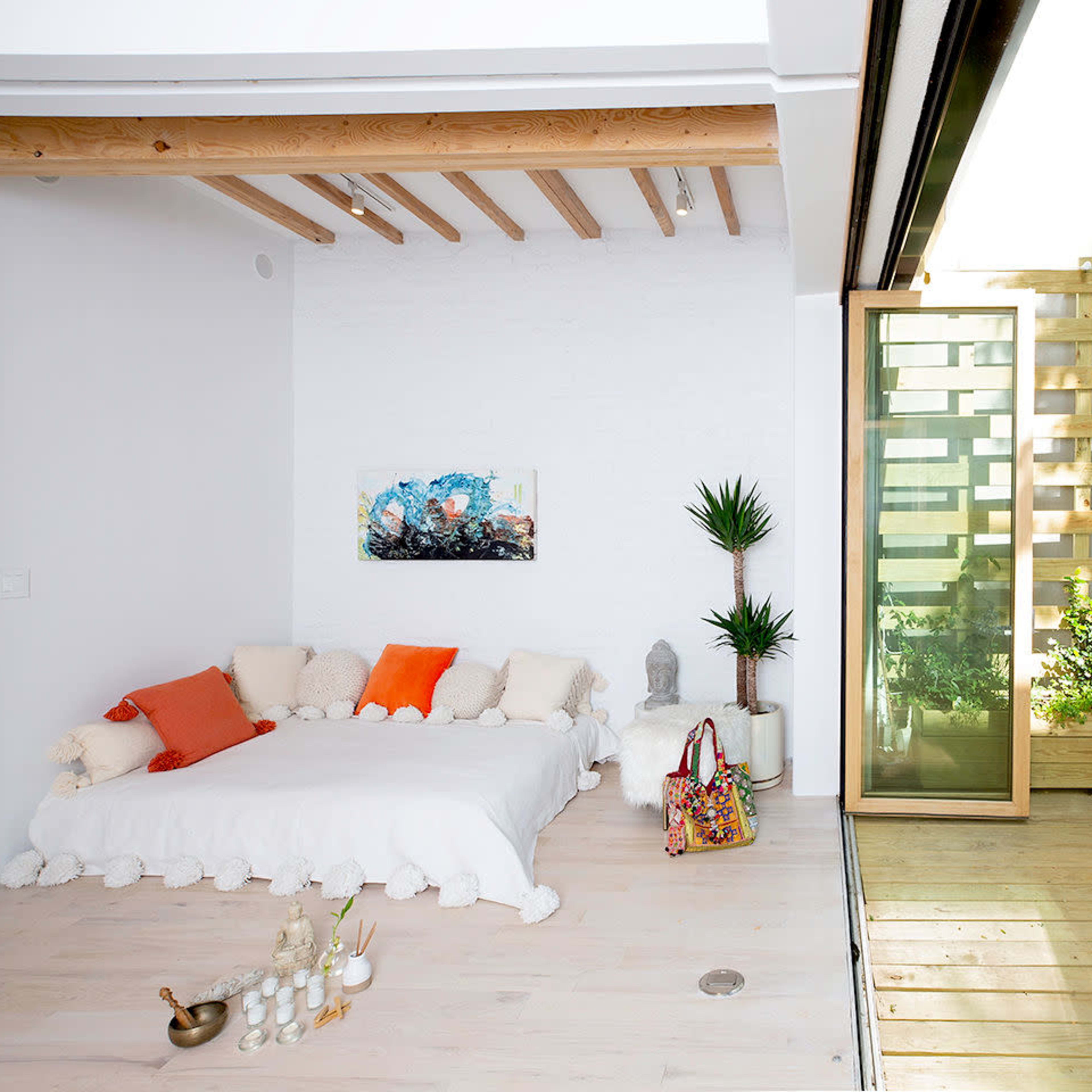 A bright, modern living space with a bed covered in light bedding on one side and a wooden deck with plants and a small dining area on the other.