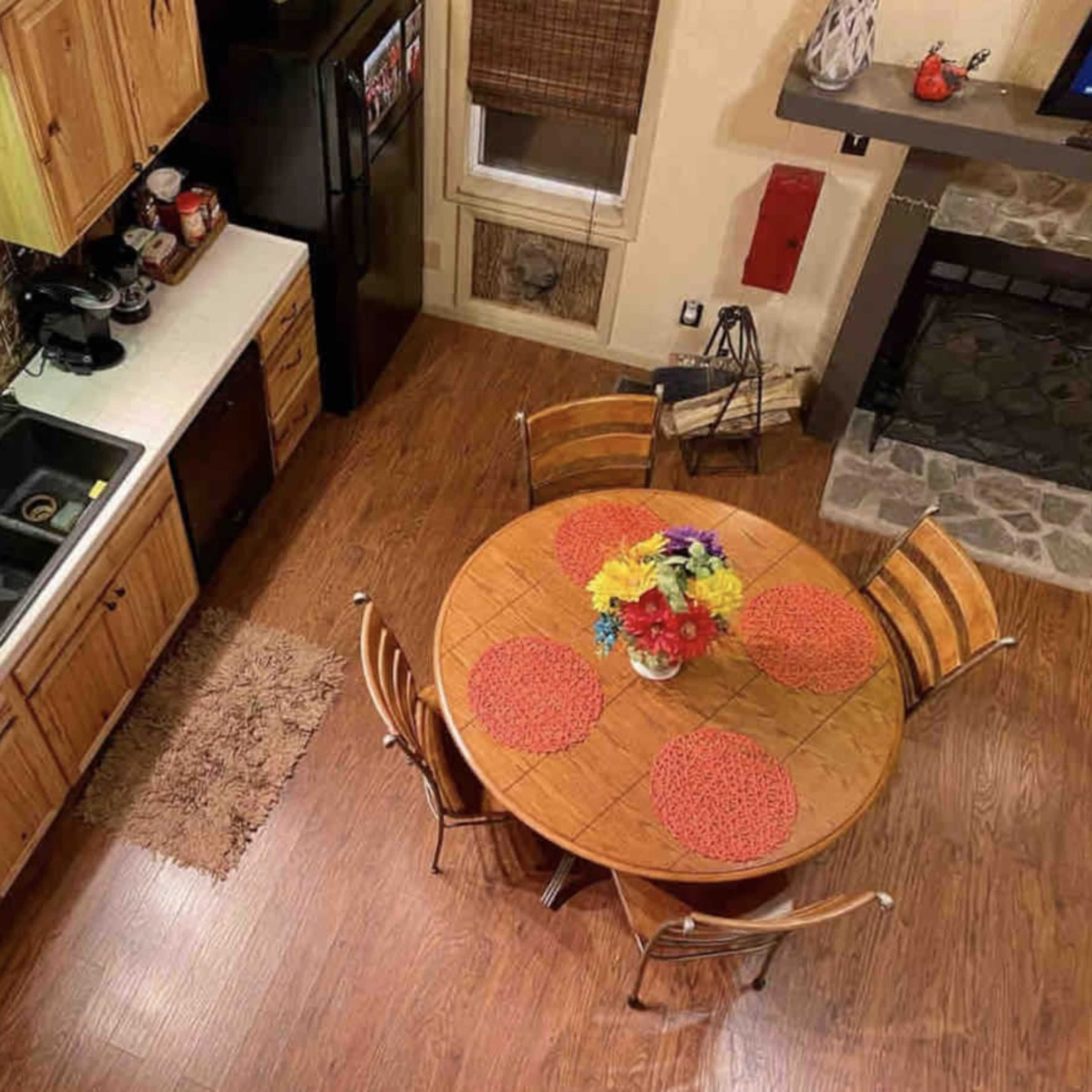 A well-organized kitchen with wooden cabinets, a round dining table set with colorful placemats and flowers, and a cozy seating area near a stone fireplace.