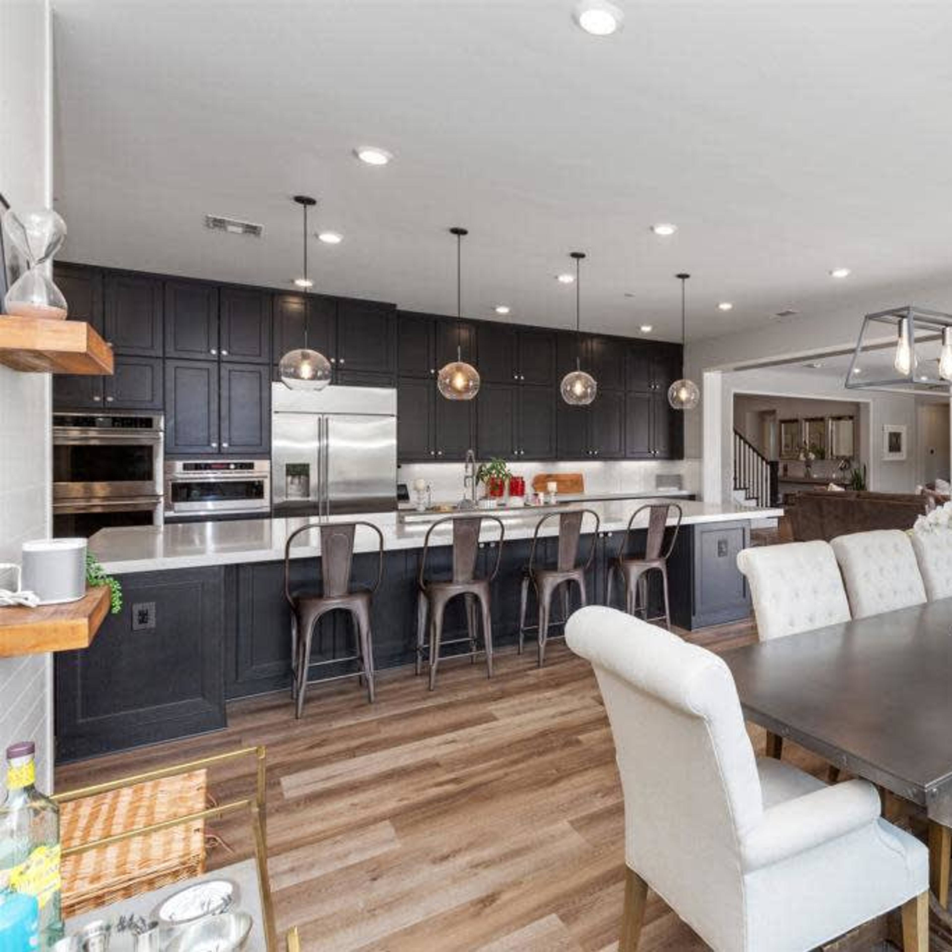 A modern kitchen and dining area feature dark cabinetry, a long dining table with upholstered chairs, and pendant lighting above the kitchen island.