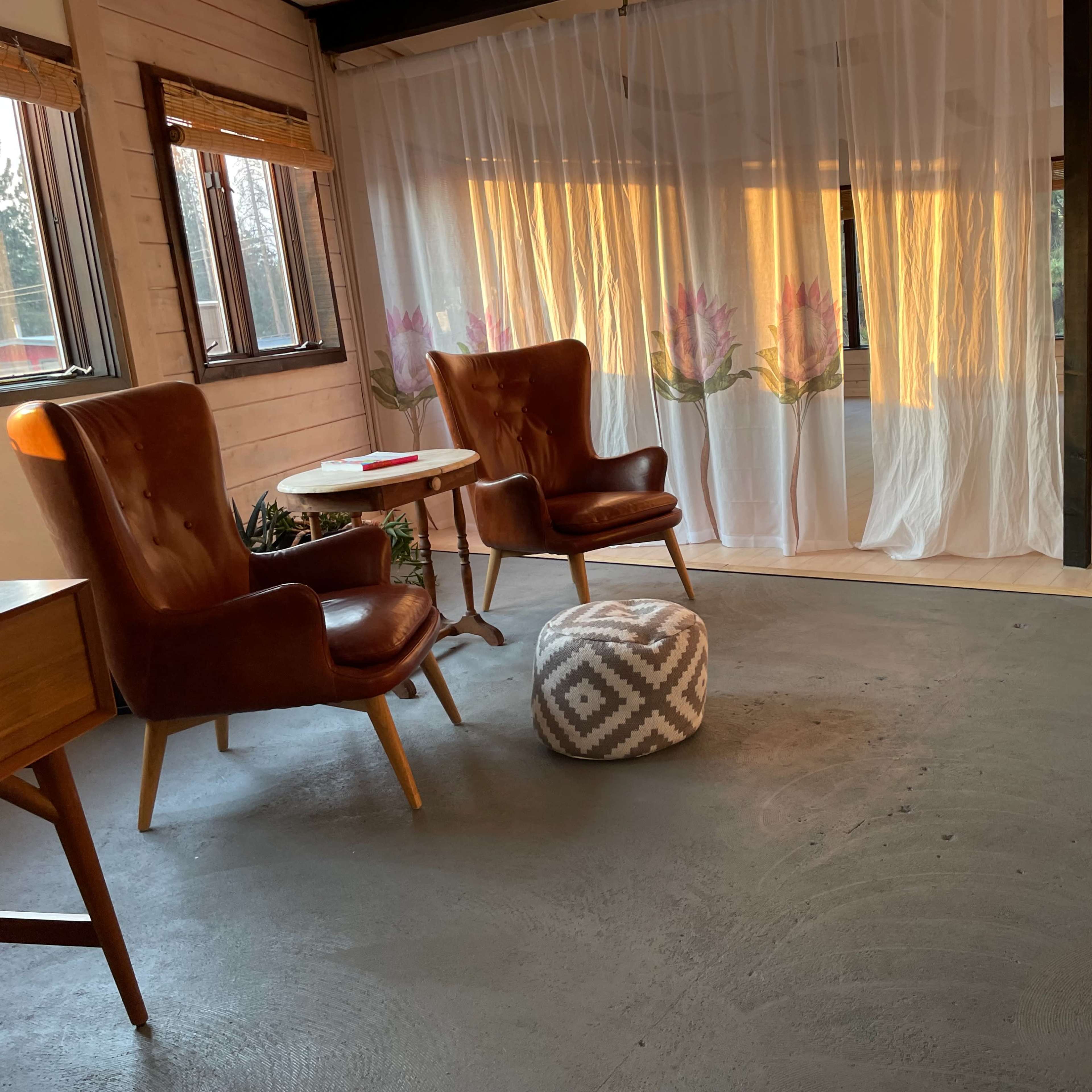 Two brown armchairs are positioned near a small circular table on a concrete floor in a room with sheer curtains featuring floral designs.