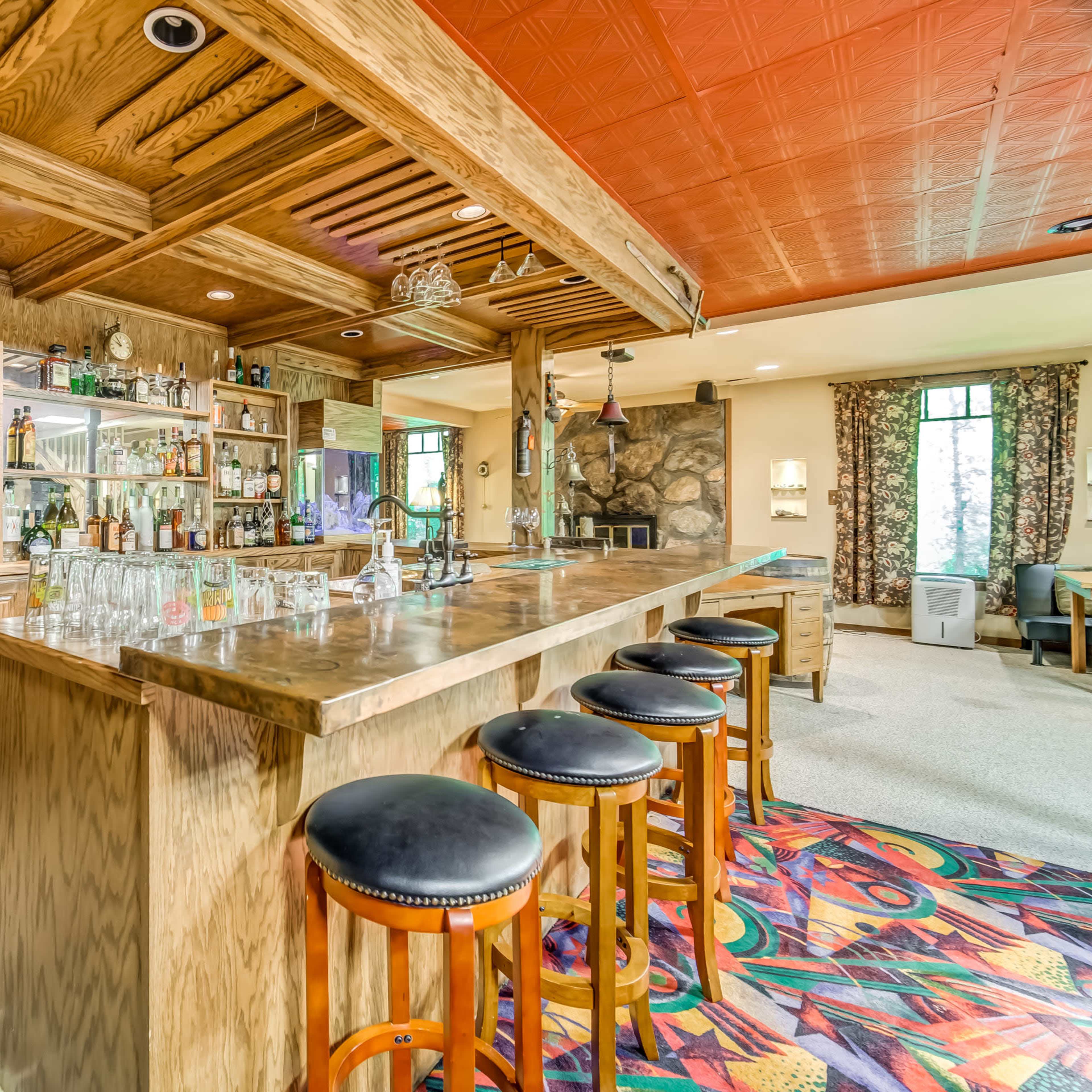 A wooden bar area features a large counter with several stools, shelves stocked with bottles, and a stone wall backdrop in a well-lit room.